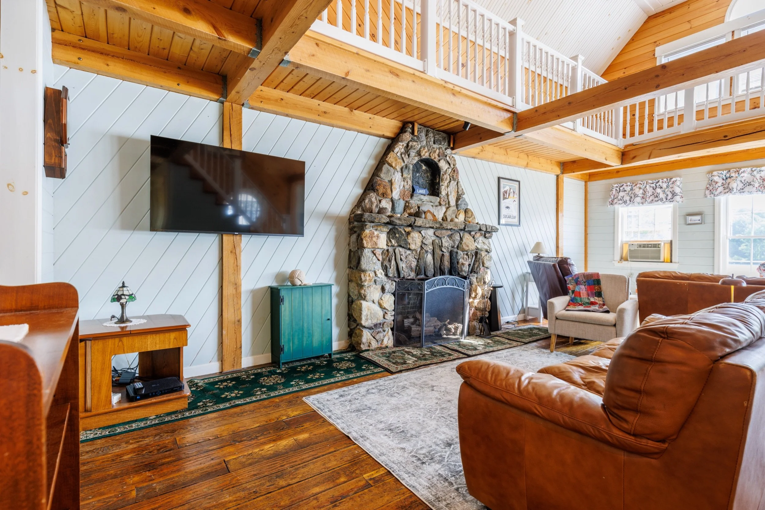 Living room with wooden beams and ceiling, stone fireplace with a sign that says 'Lake', flat screen TV, armchair with a blanket, leather couch, floral curtains, window air conditioner, and various decorative items.