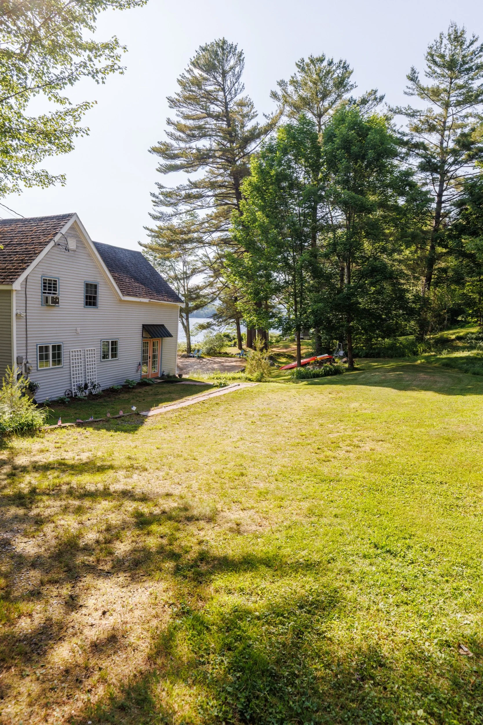 A backyard with a well-kept grassy lawn, tall trees, a white house with a sloped roof, and a view of a body of water in the background on a sunny day.