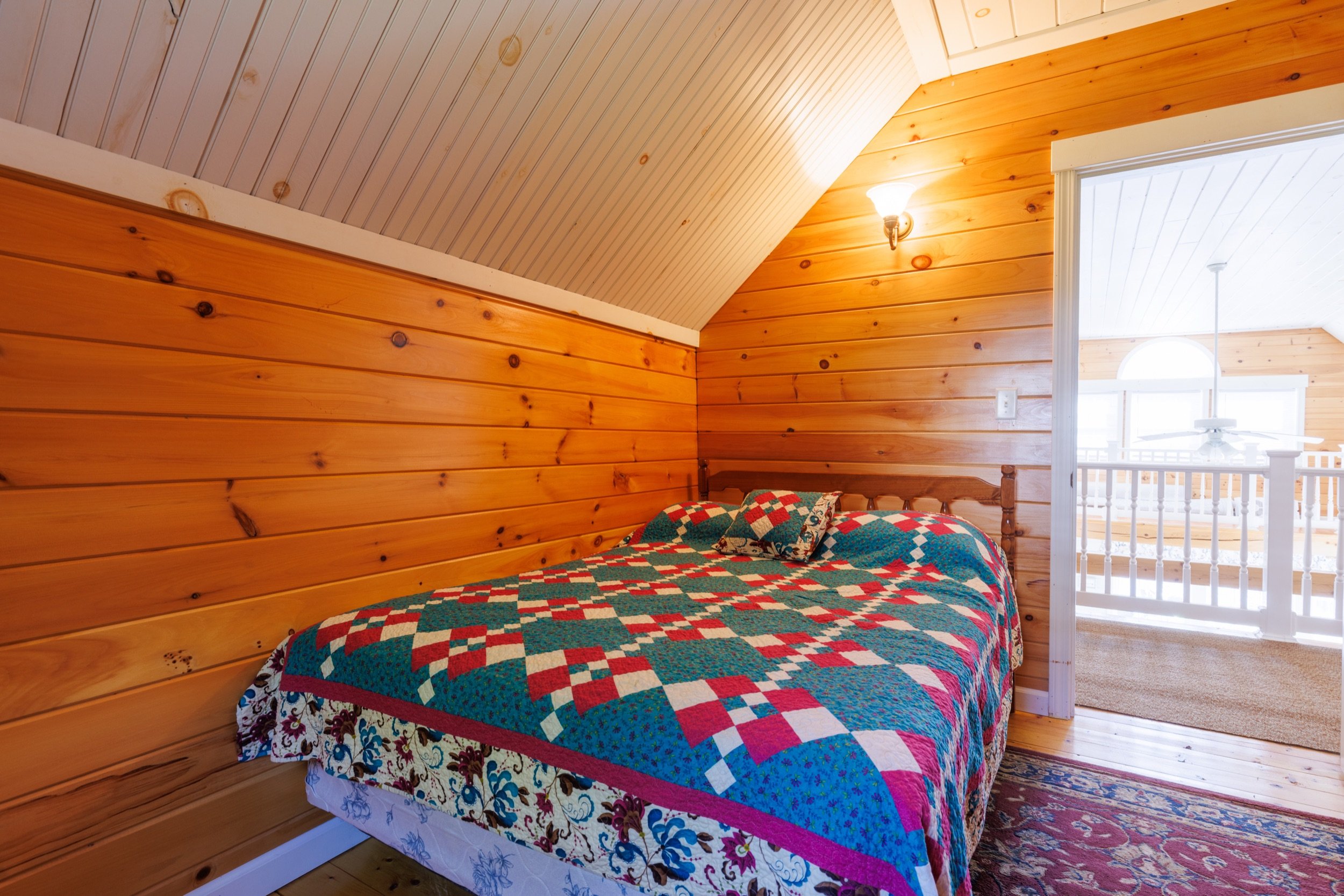 A bedroom with wooden paneling on the walls and ceiling, a bed with a colorful patchwork quilt, and a partial view of a balcony with a railing and arched window.