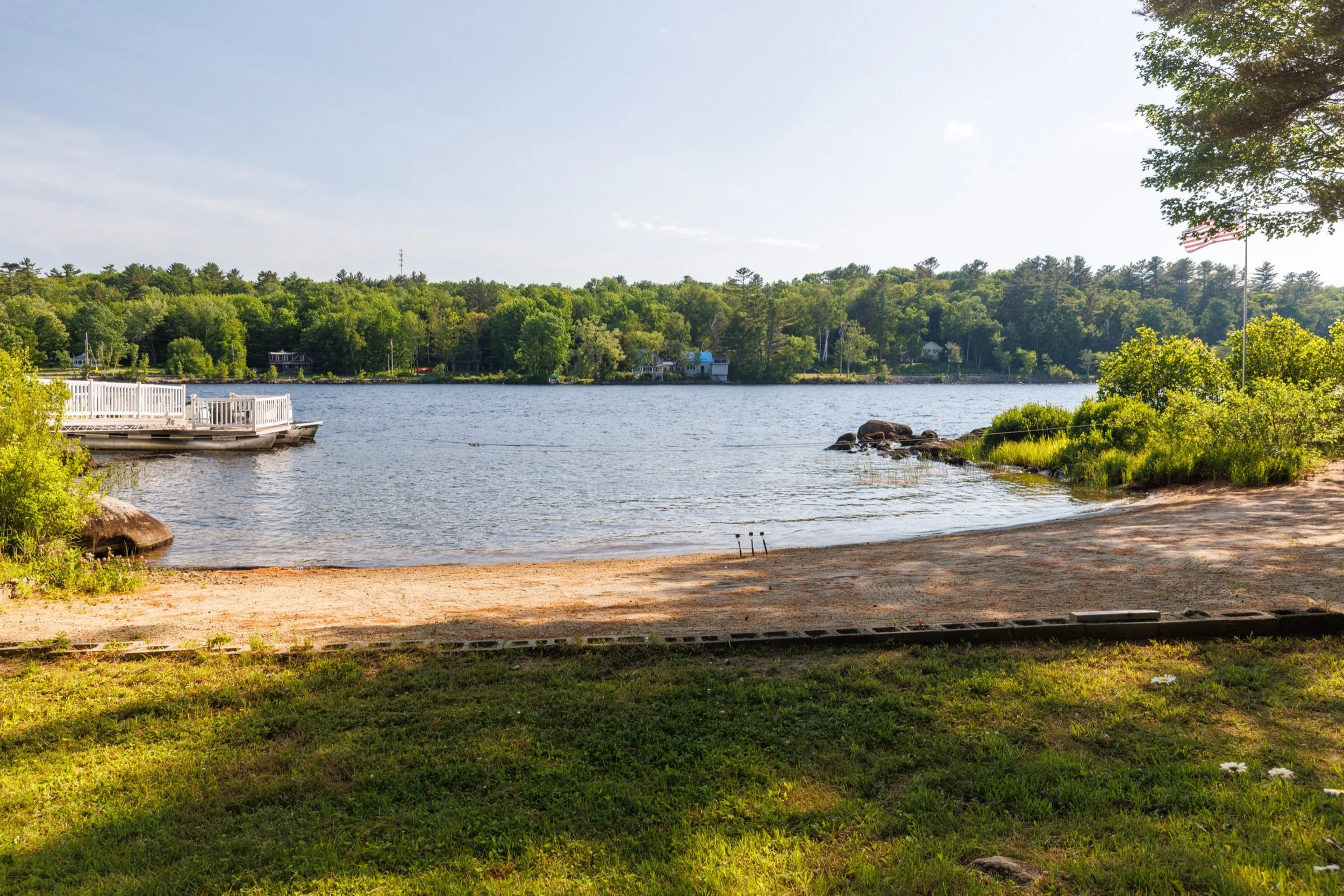 A peaceful lakeside scene with a sandy beach, green trees, a dock on the water, and houses across the lake, with a partly cloudy sky and sunlight reflecting on the water.