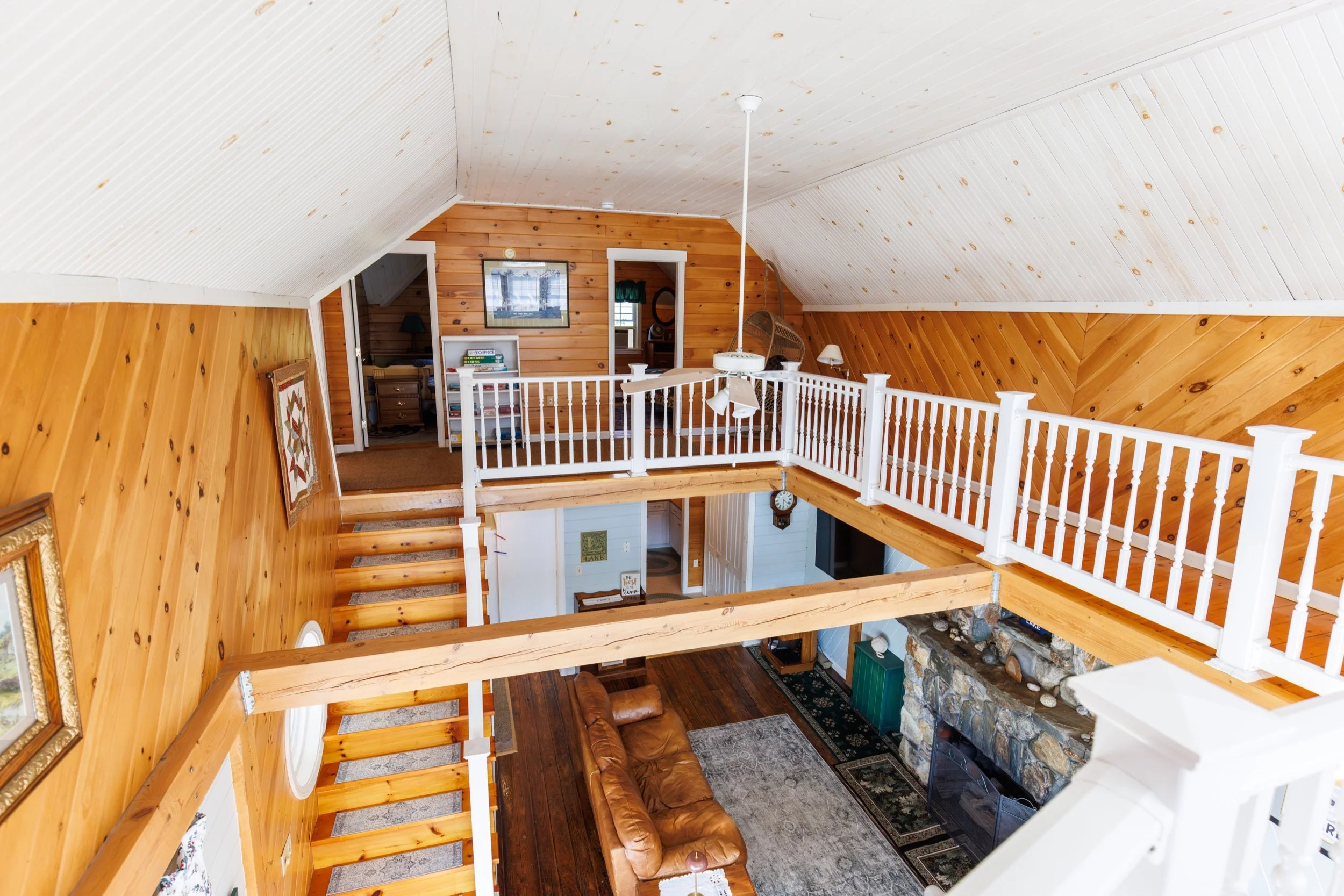 Interior view of a two-story wooden house with a living room and an upper loft area. The house has wood-paneled walls and ceiling, with white railings around the loft. Furniture includes a leather sofa, various lamps, a coffee table, and decorative w