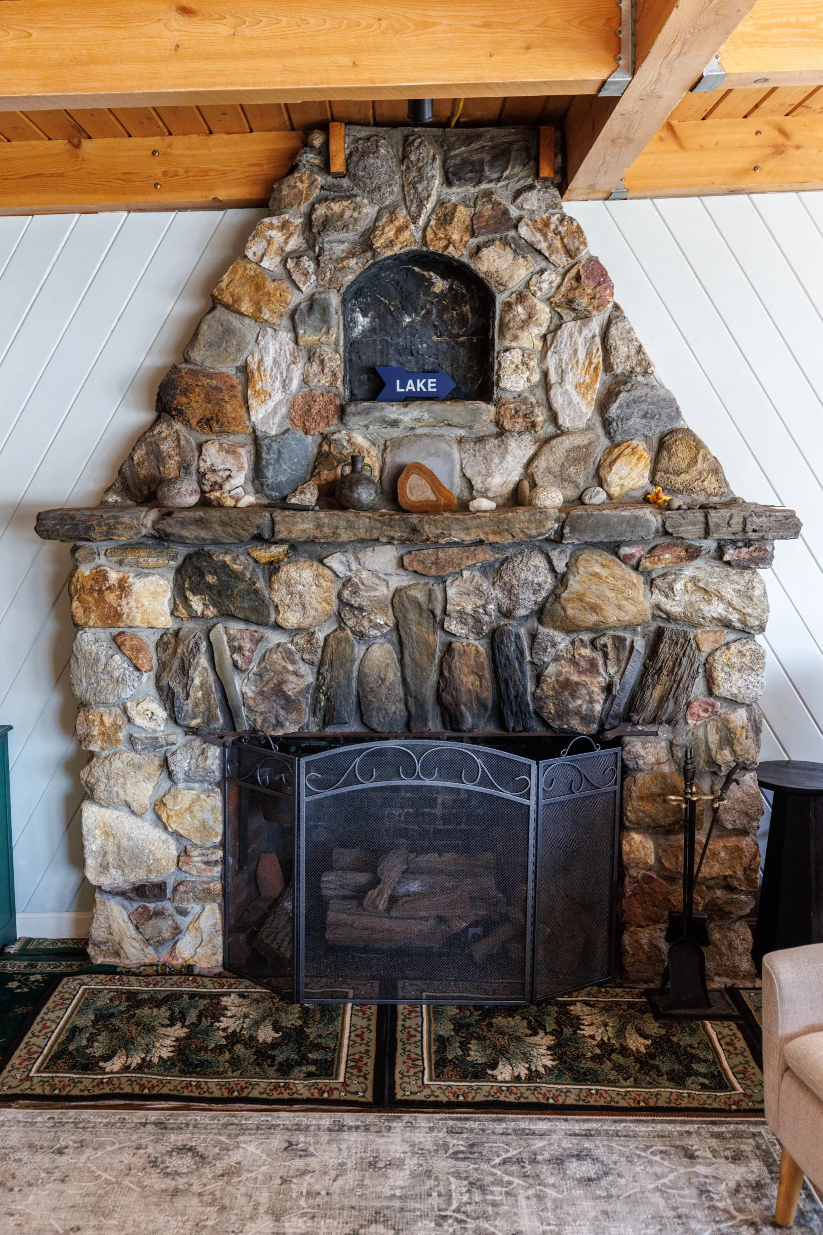Stone fireplace with a black metal fire guard, a small decorative bottle, and a sign that says "Lake" inside the hearth, wooden beam ceiling, white wood wall paneling, patterned area rug, and part of a beige armchair visible on the right.