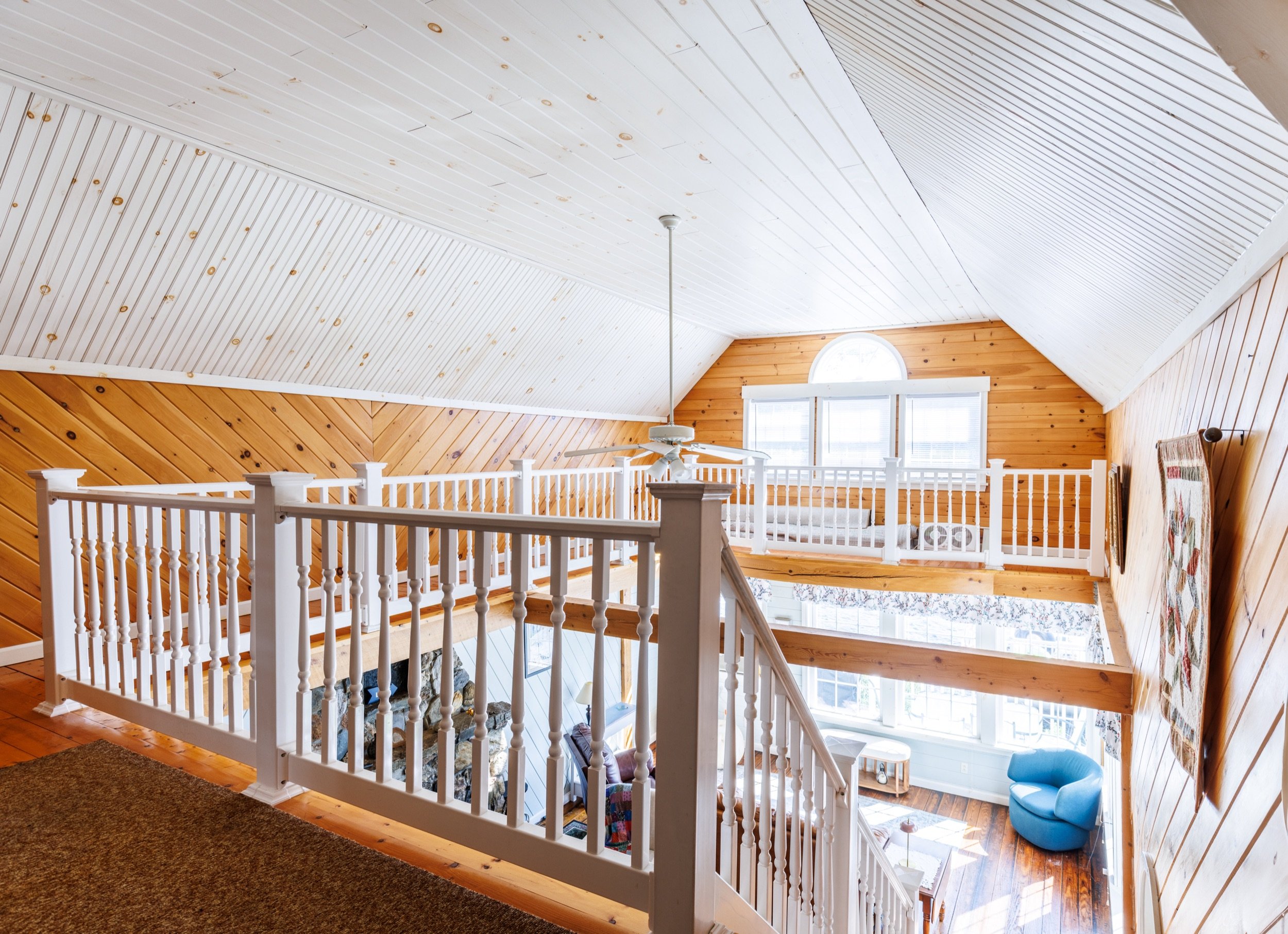 Interior view of a spacious room with a vaulted ceiling, finished with white painted wood planks and natural wood paneling, large windows, a ceiling fan, and a cozy seating area with a blue chair.