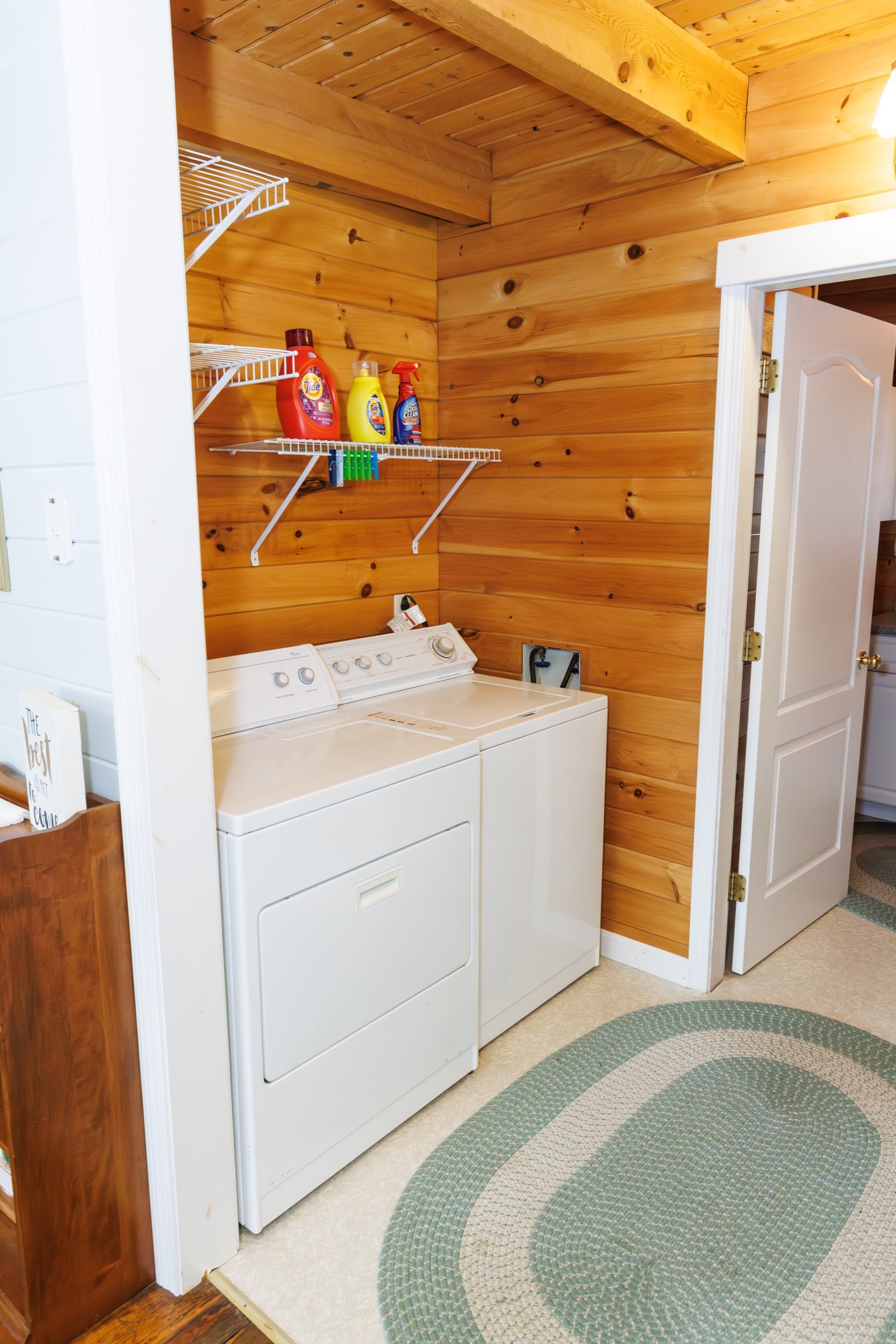 Laundry area with wooden wall paneling, a white top-loading washer and dryer, a circular patterned rug on the floor, and a white wire shelf with laundry supplies.