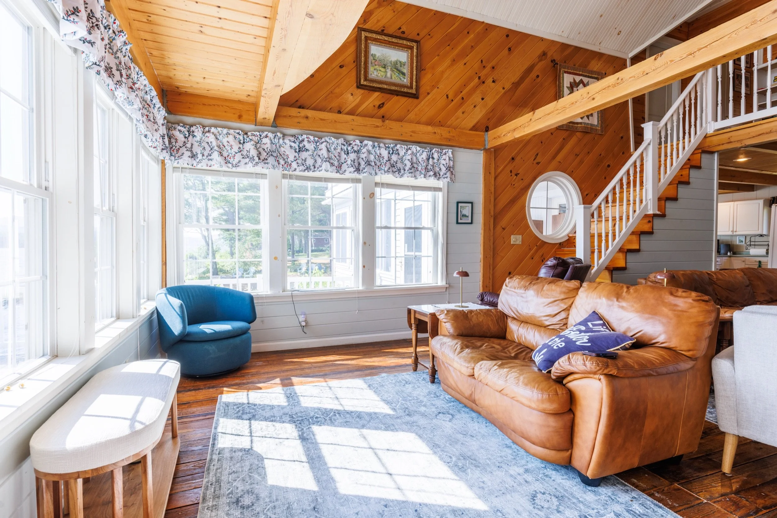 Living room with large windows, white walls, wooden beams, and ceiling, brown leather sofa, blue chair, white bench, and patterned curtains.