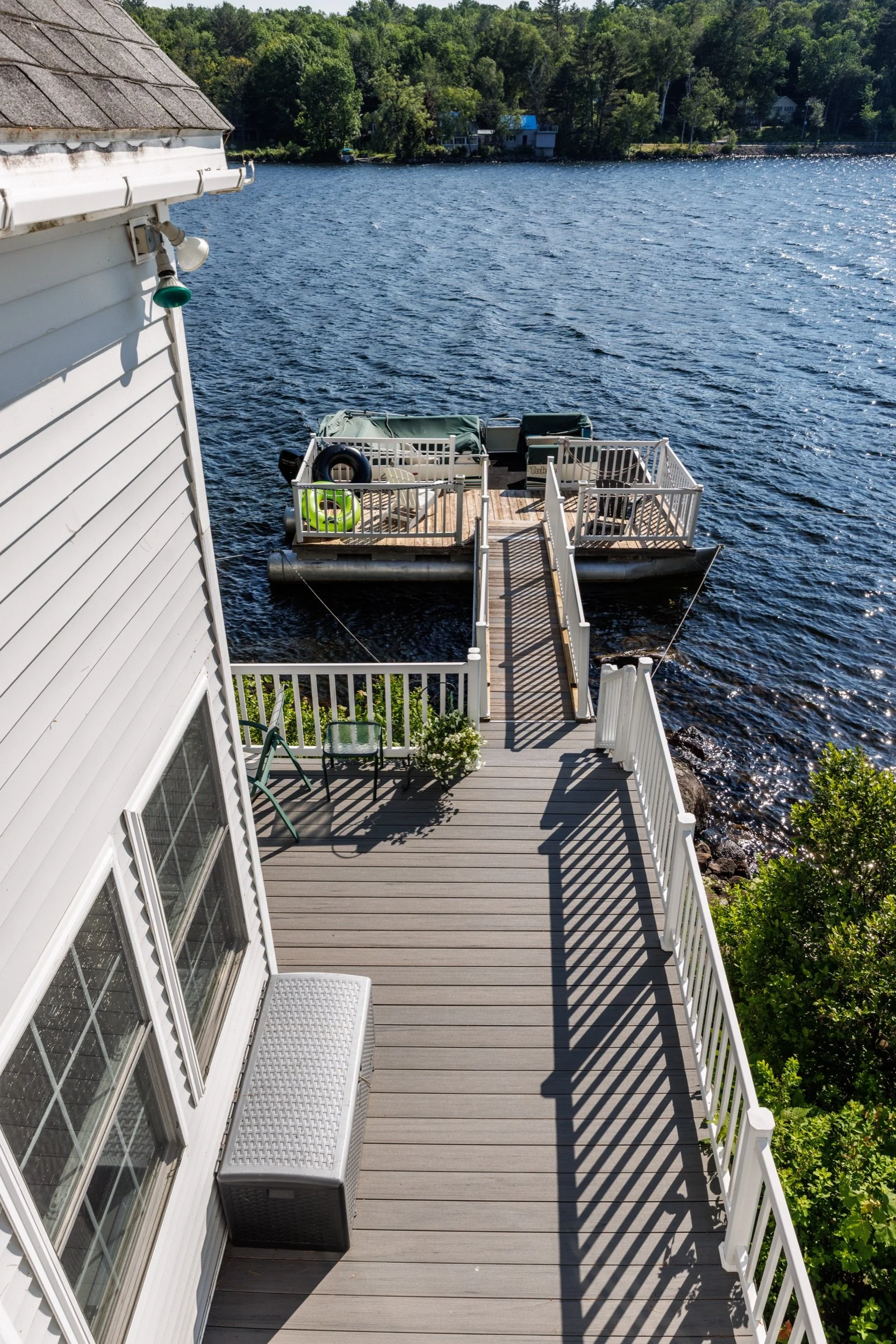 A wooden deck with white railings overlooking a lake, with chairs and outdoor furniture, partially shaded by a house, and surrounded by trees.