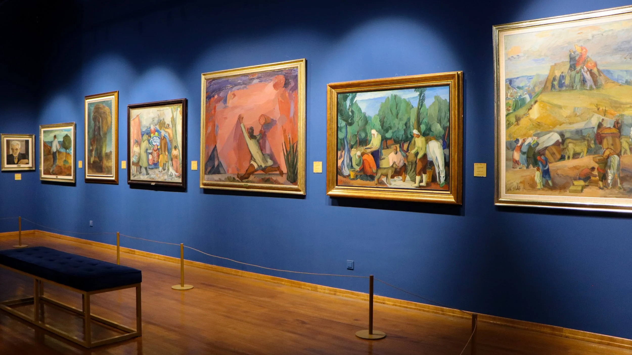 Paintings in an art gallery displayed on a blue wall, with a bench in front.