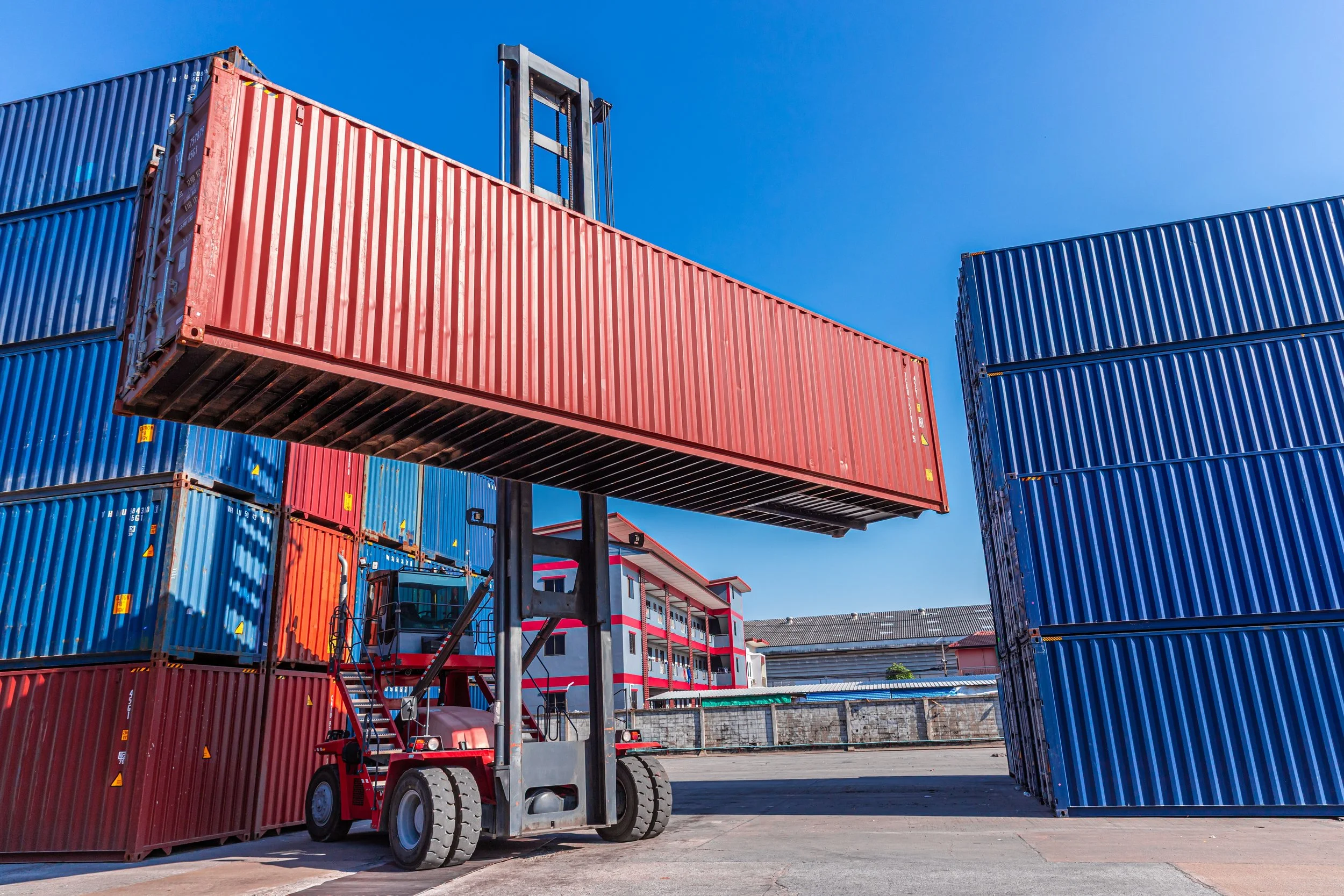 A forklift lifting a red shipping container in a port, with additional blue containers stacked nearby and a building with red accents in the background.