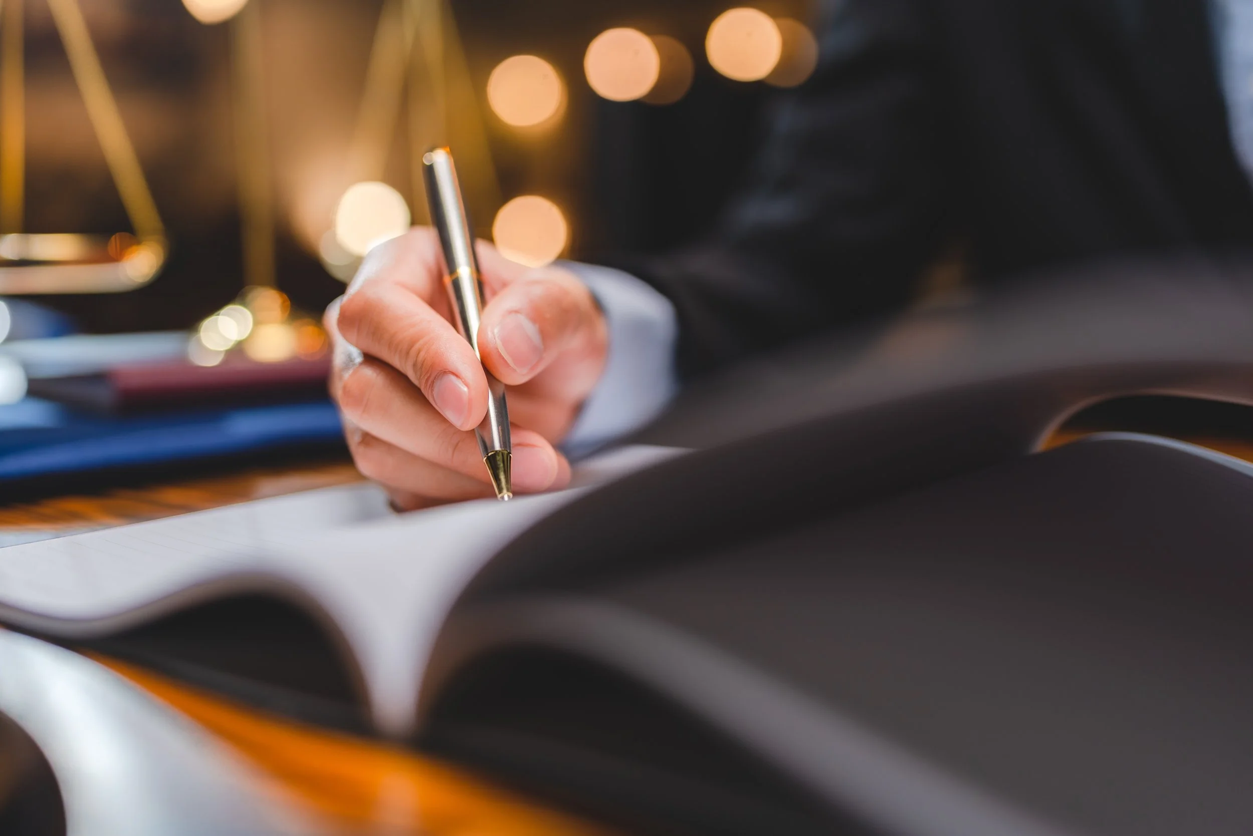 Close-up of a person's hand writing in a notebook with a pen, with background blurred lights.