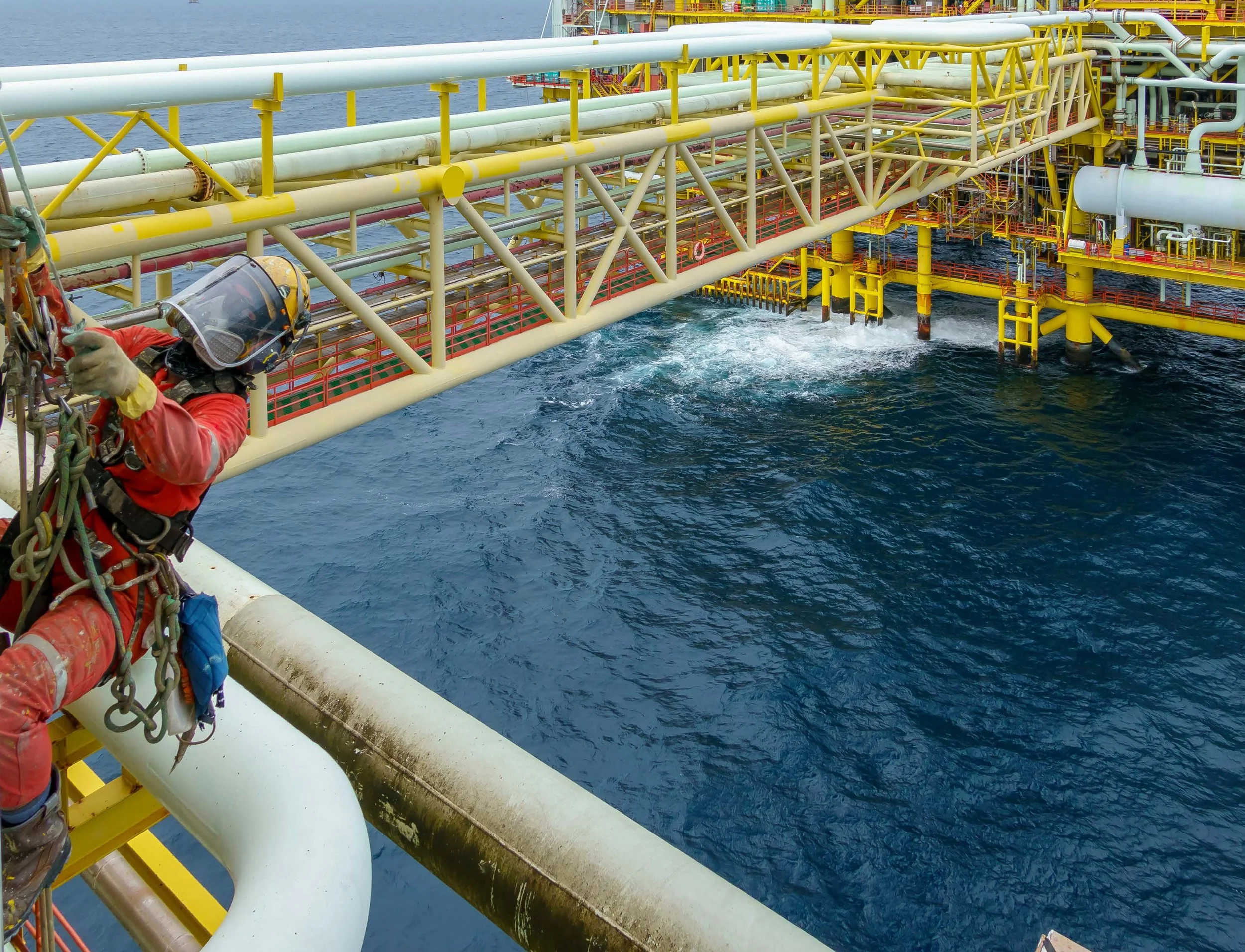 Offshore oil platform with yellow and white pipelines and structures, person in safety gear securing themselves to the platform railing, ocean in the background.