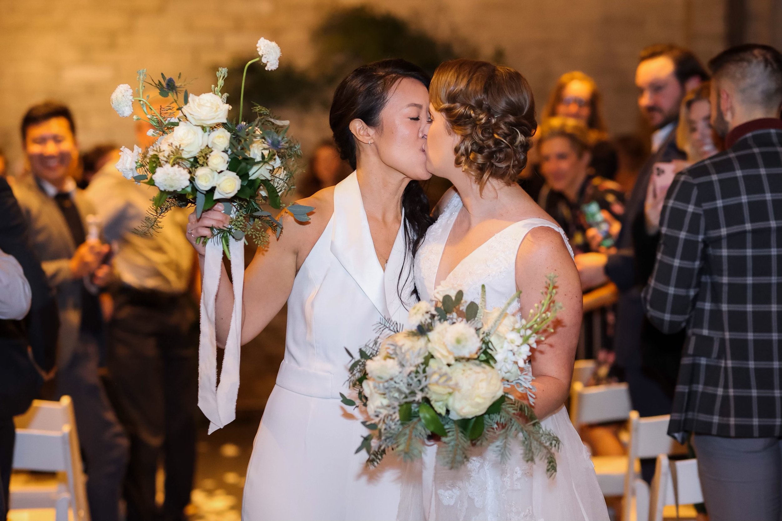 Two women kiss at a wedding ceremony, holding bouquets of white flowers, with guests in the background - AXIS, Seattle WA, Gordon Duo Weddings