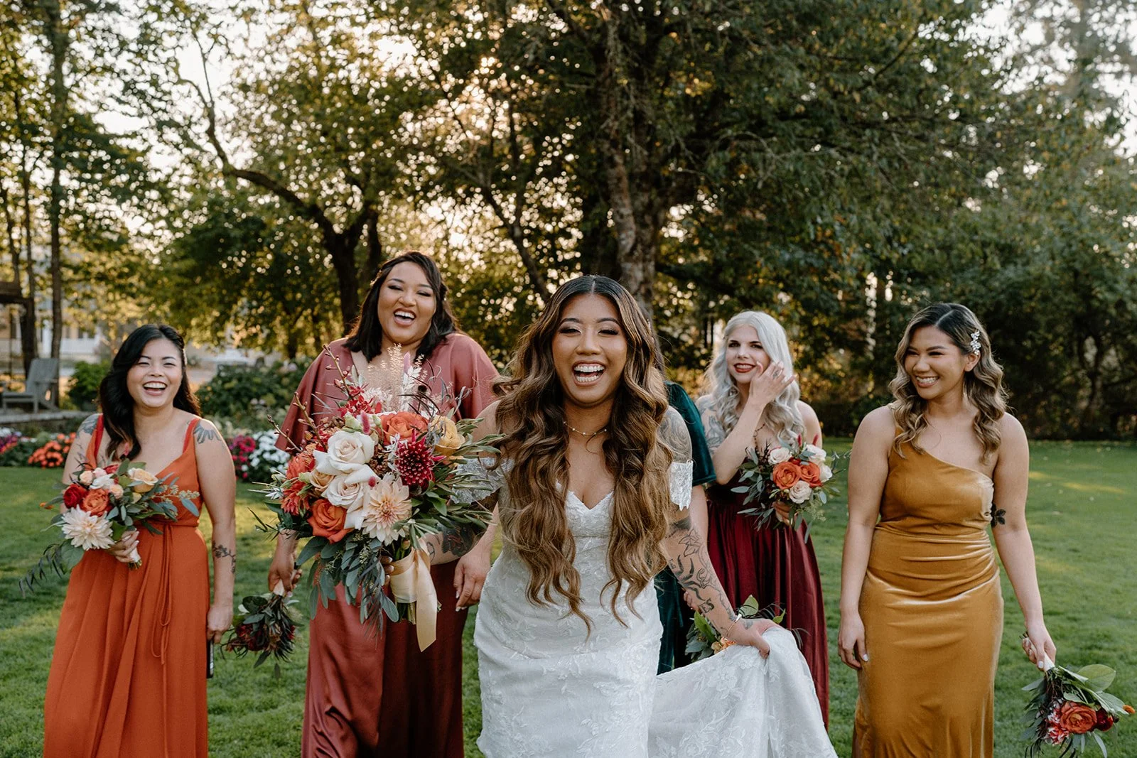 A group of five women, including a bride in a white wedding gown and four bridesmaids in colorful dresses, walking outdoors in a park or garden setting with trees and flowers in the background, all smiling and holding bouquets - Gordon Duo Weddings
