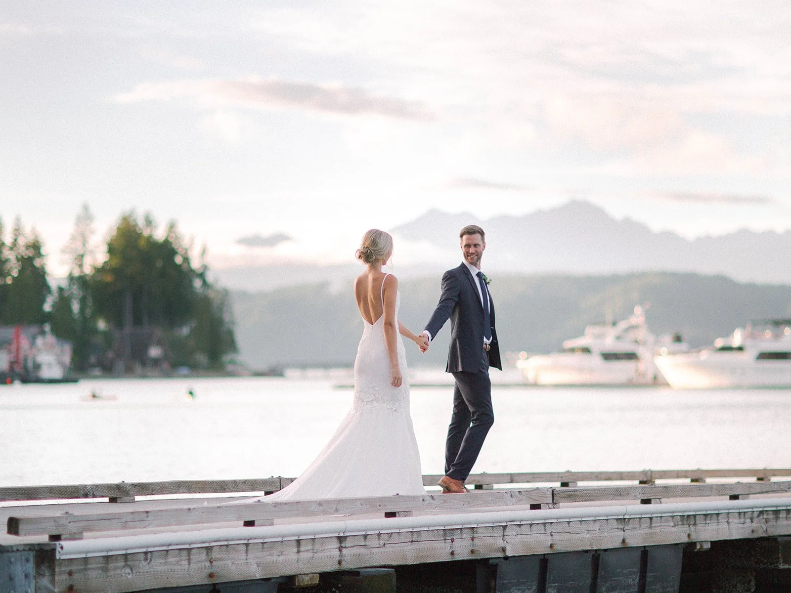 A newlywed couple holding hands on a dock by a body of water during sunset, with boats and mountainous landscape in the background - Alderbrook Resort & Spa, Union WA — Gordon Duo Weddings, photo by Rachel Haylie