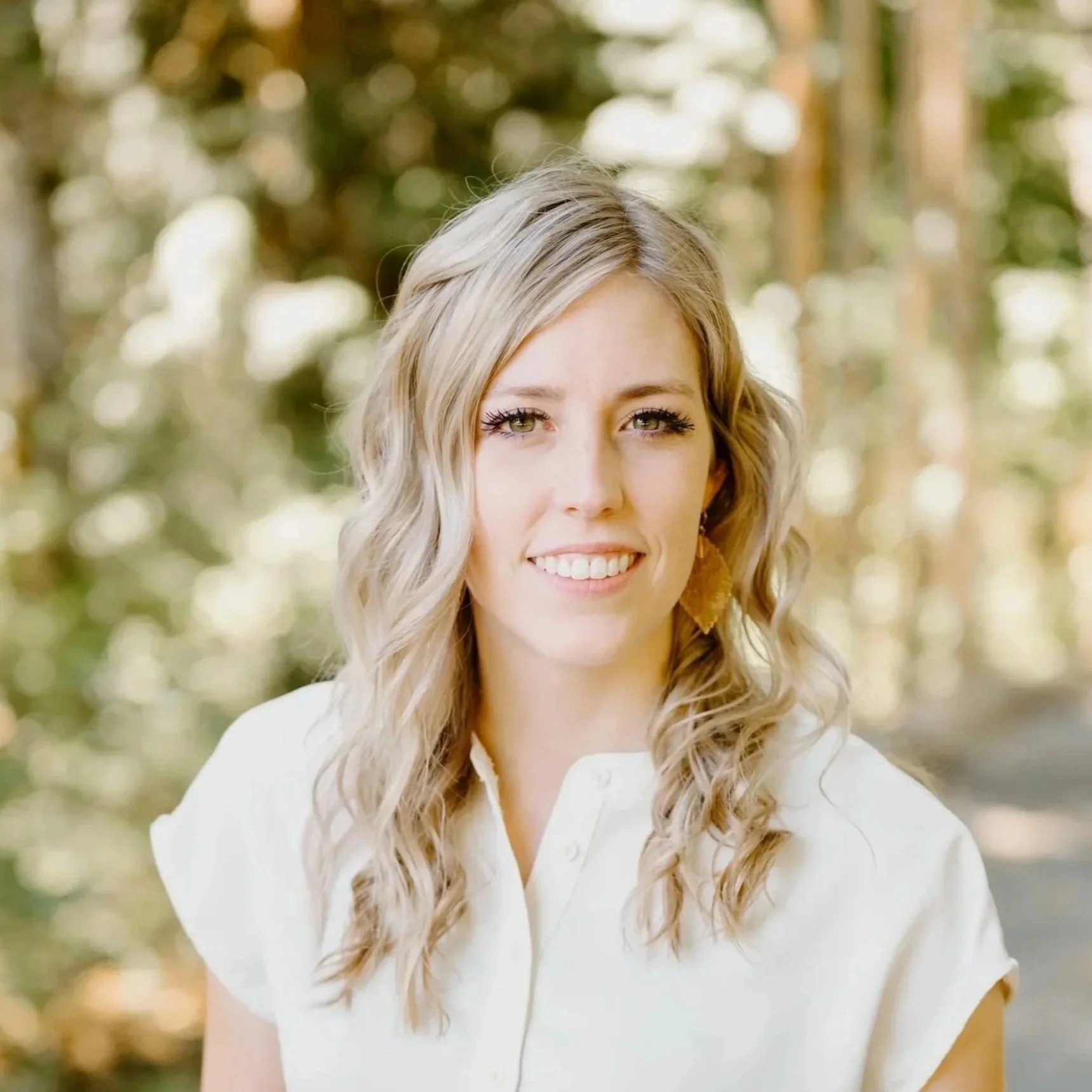 Close-up of a young woman with blonde, curly hair smiling outdoors with a blurred background of trees and sunlight. Lead Coordinator, Gordon Duo Weddings