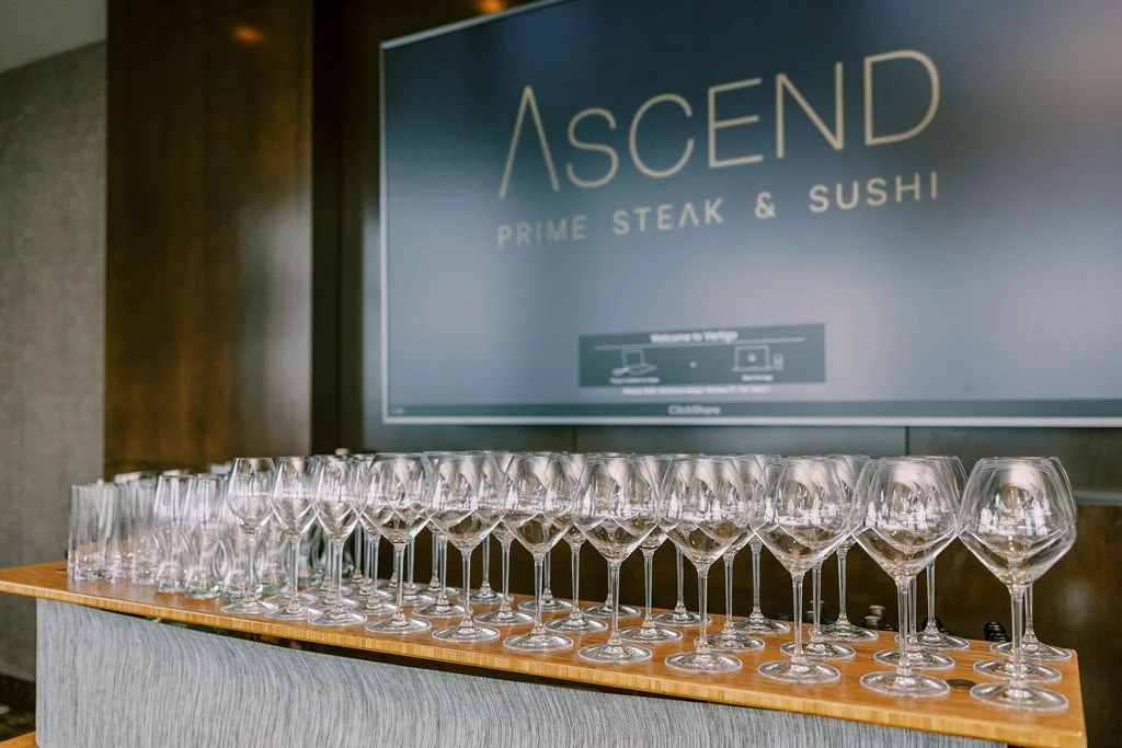 A row of empty wine glasses on a wooden bar counter in front of a sign reading 'ASCEND PRIME STEAK & SUSHI' -  W Bellevue and ASCEND, Bellevue WA — Gordon Duo Weddings, photo by Vivi Lin