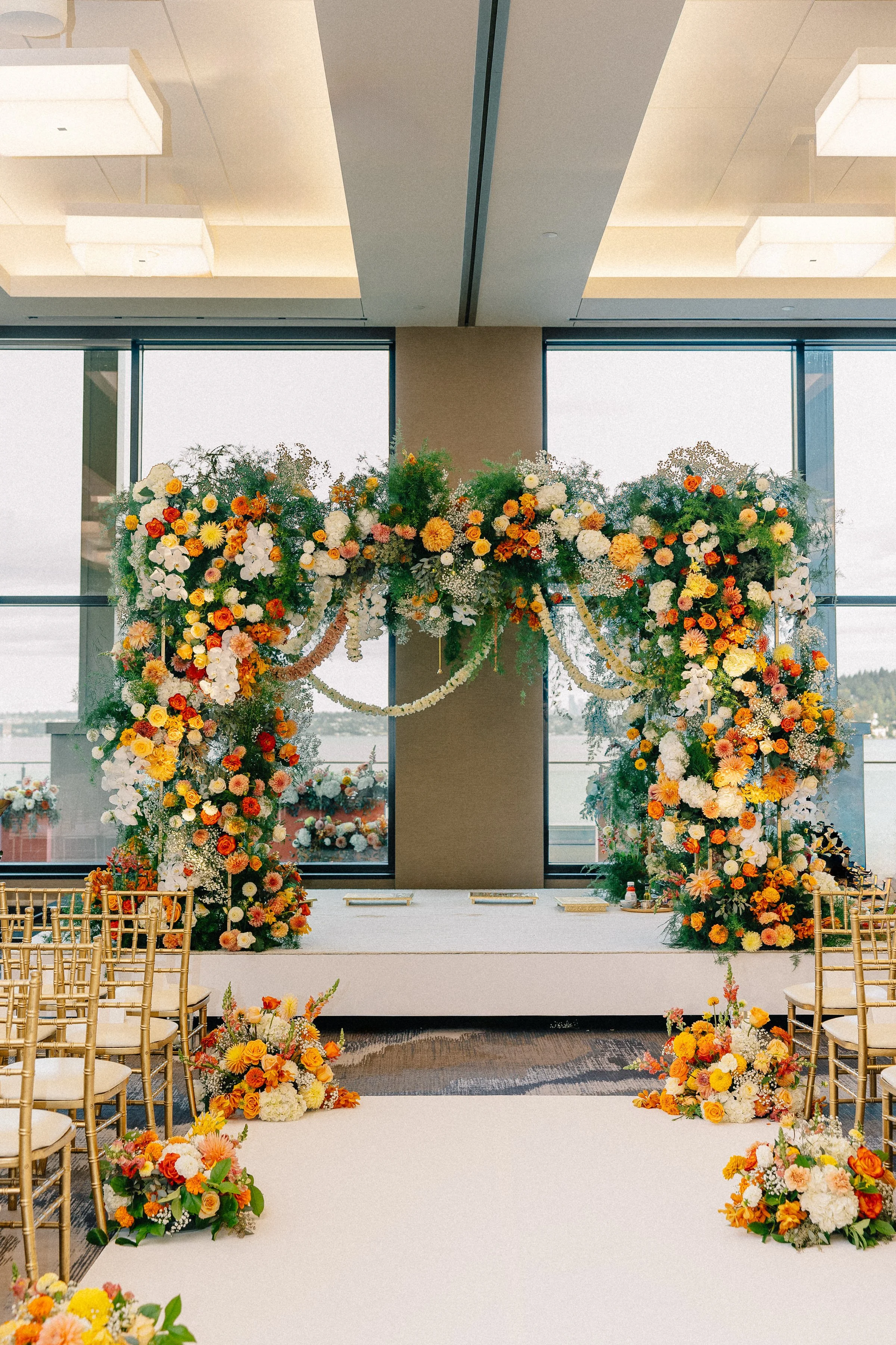 A decorated ceremony arch with a floral arrangement of white, orange, and yellow flowers, set inside a modern building with large windows and chairs arranged on each side - Hyatt Lake Washington, Renton WA — Gordon Duo Weddings, photo by Hannah Hix