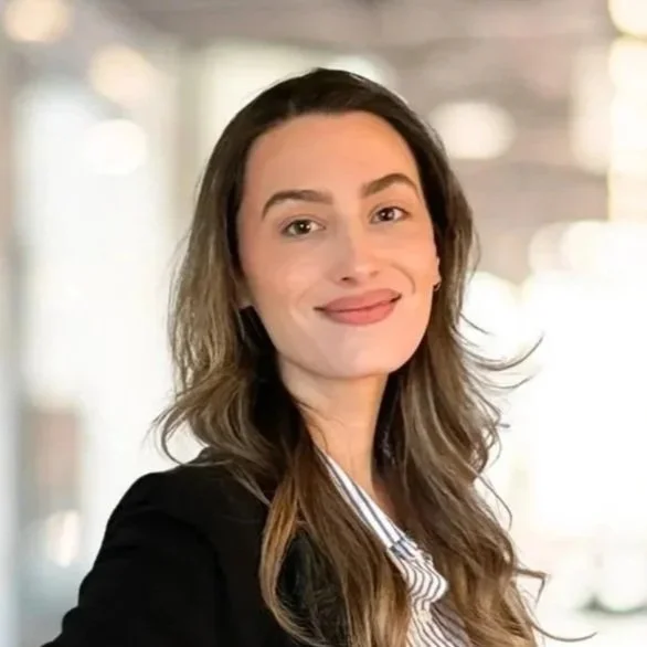 A woman with long wavy hair, wearing a blazer and striped shirt, smiling in a well-lit indoor setting. Assistant Coordinator, Gordon Duo Weddings
