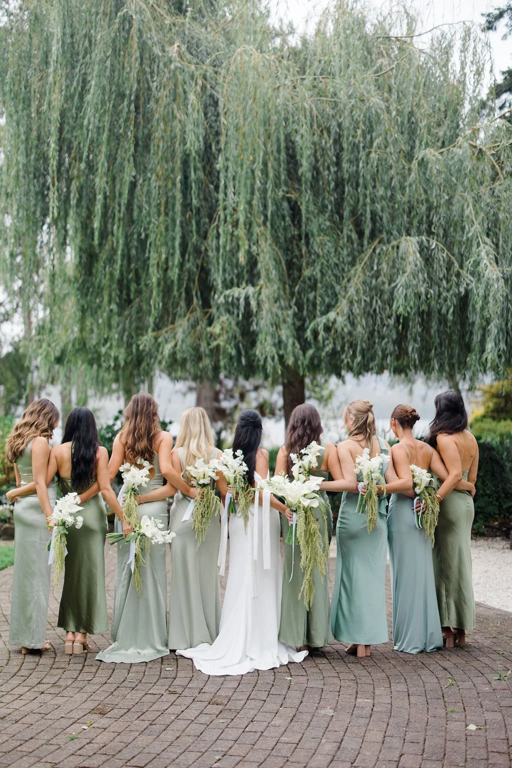 A bridal party standing in a row outdoors, each woman holding a white bouquet, with a large leafy tree overhead and greenery in the background - Kiana Lodge, Pulsbo WA — Gordon Duo Weddings, photo by Claire Forrest