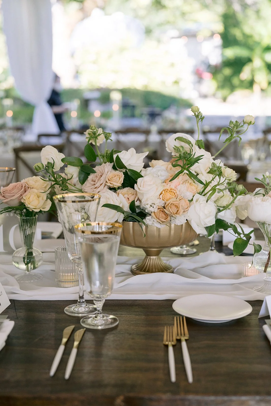 Wedding reception table decorated with a large centerpiece of white and blush roses, surrounded by glassware, gold-rimmed champagne flutes, candles, white plates, and gold and white flatware, set outdoors with greenery in the background  - Gordon Duo