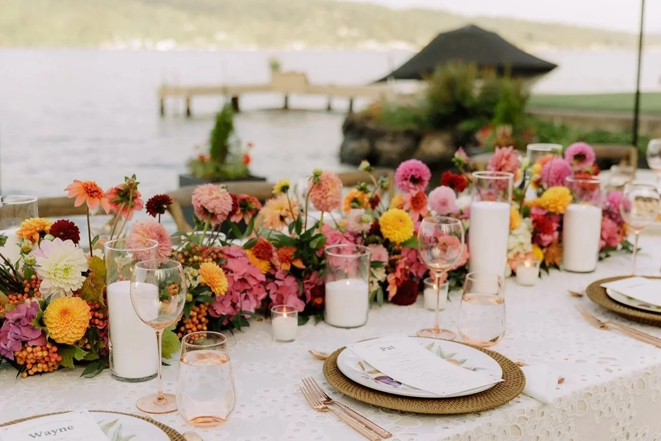 A decorated outdoor dining table with pink, yellow, and orange flowers, white candles, and glassware near a body of water with a wooden dock and pavilion in the background - Home Wedding , Seattle WA — Gordon Duo Weddings, photo by Jessica Wood