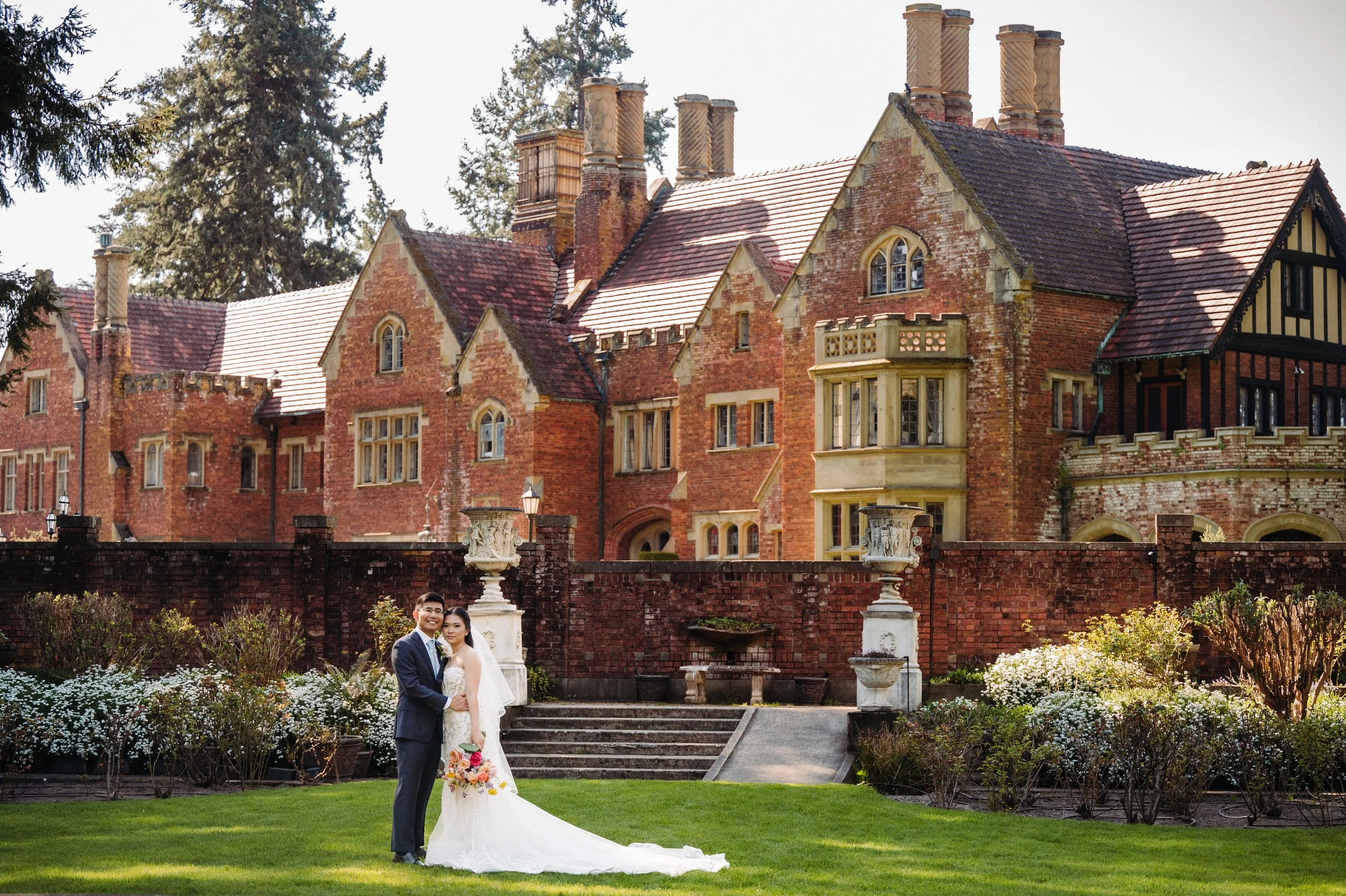 A bride and groom in wedding attire standing on a garden lawn in front of a brick building with multiple chimneys. The bride is holding a bouquet and the couple is smiling, with lush greenery and flowers in the surrounding garden.