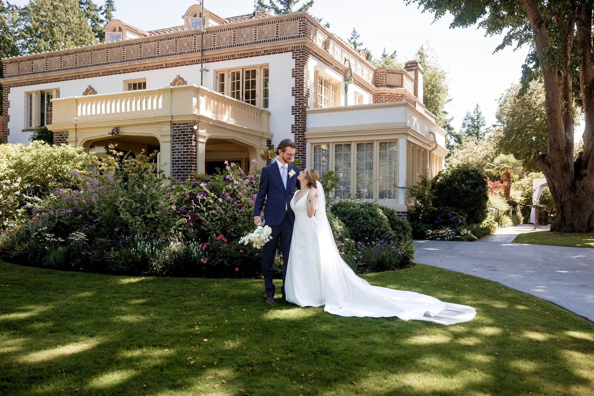 A bride and groom standing on a lawn in front of a large historic brick house with white trim, surrounded by colorful flowers and greenery, during a sunny day - Lairmont Manor, Bellingham WA — Gordon Duo Weddings, photo by Bear Beau
