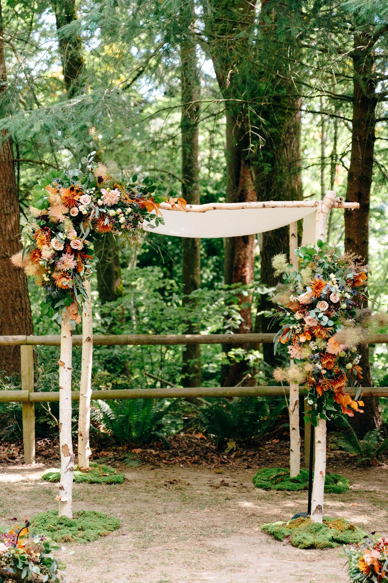 A wedding arch decorated with flowers and greenery in a forest setting, with trees and foliage in the background. The Lodge at St Edward Park, Kenmore WA — Gordon Duo Weddings, photo by Cameron Zegers