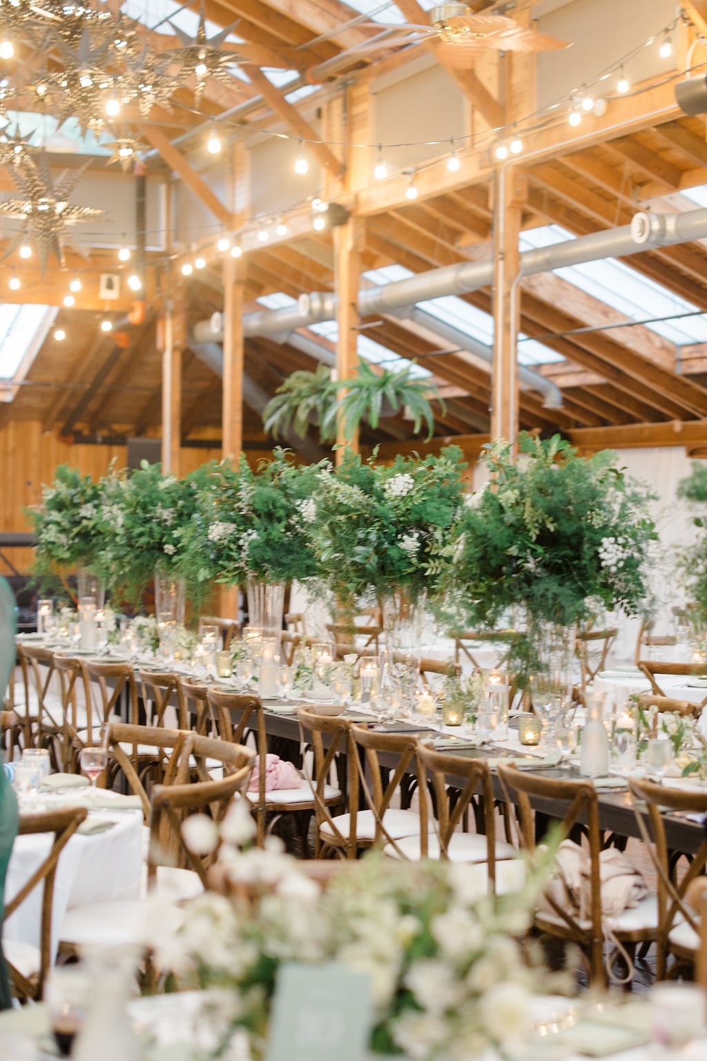 Indoor event space decorated with string lights, large floral arrangements, and wooden chairs at a long dining table - Kiana Lodge, Pulsbo WA — Gordon Duo Weddings, photo by Claire Forrest