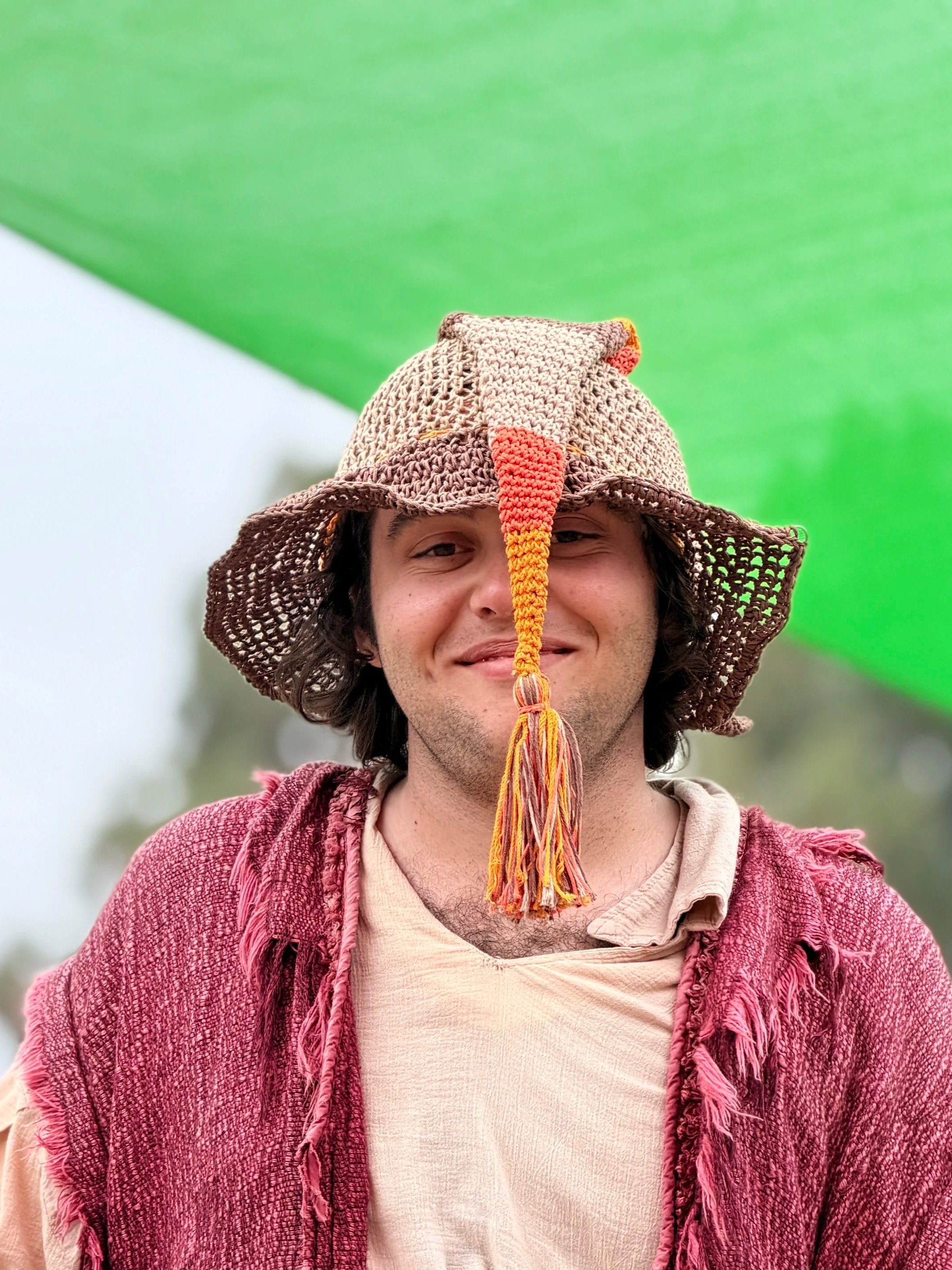 A person wearing a crocheted hat with a long tail, a beige shirt, and a red textured jacket, smiling outdoors at a renaissance faire.