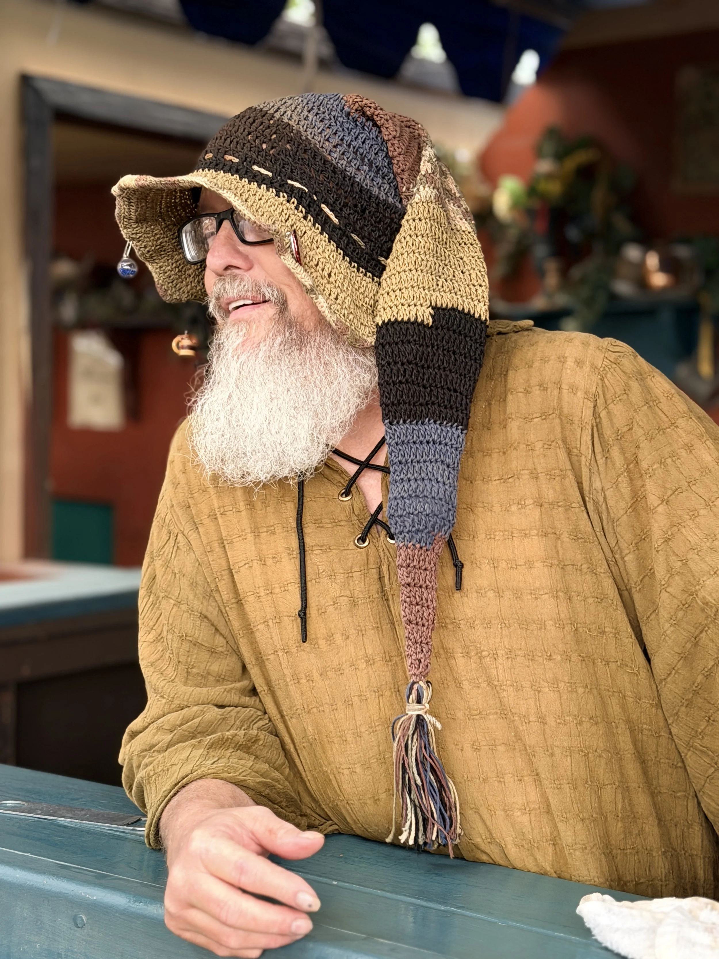 A man with a long gray beard wearing glasses is smiling while working at an ale stand at a Renaissance Faire.  He is wearing a colorful crocheted hat with a brown shirt. The background includes shelves with decorative items and plants.