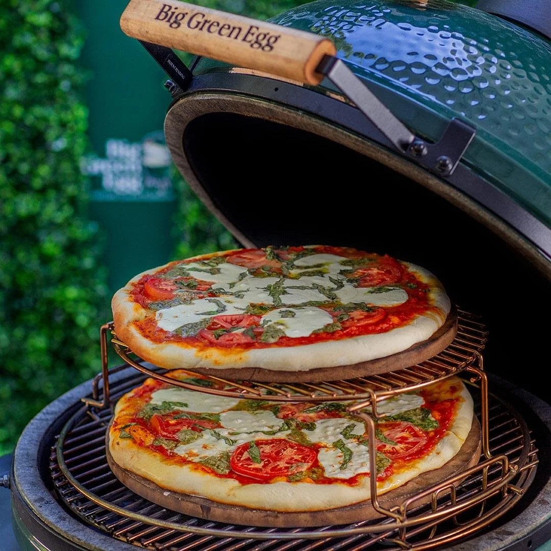 Two pizzas inside a green outdoor grill with a wooden handle labeled 'Big Green Egg', cheese, tomato, and basil toppings, on metal racks for cooking.