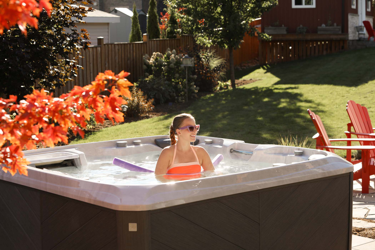 A woman sitting in an outdoor hot tub on a sunny day in a backyard with trees and shrubs, red Adirondack chairs, and a wooden fence.