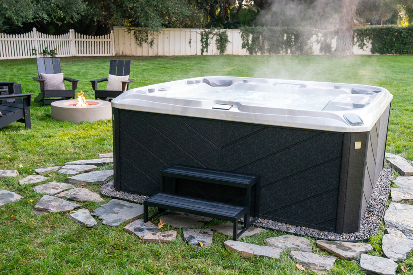 Outdoor backyard with a hot tub, fire pit, chairs with pillows, and a white fence in the background.