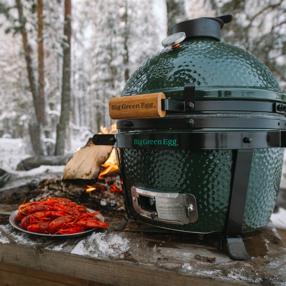 Green Big Green Egg grill on outdoor wooden table, cooking lobster, with snow and trees in the background.