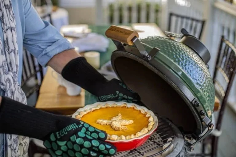 Person wearing a blue shirt and black oven mitts opening a green ceramic grill to serve a pumpkin pie with decorative whipped topping.