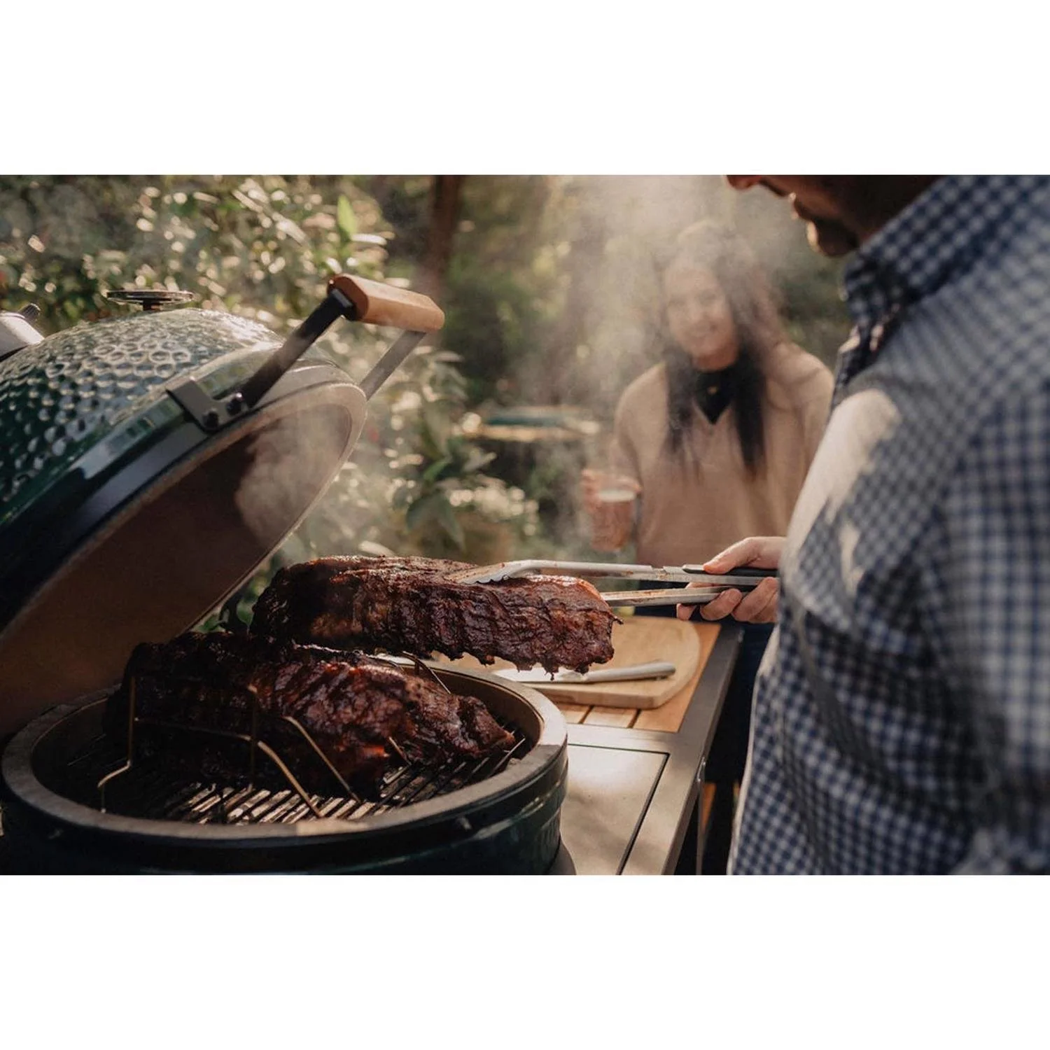 People cooking barbecue outdoors with a green smoker grill and smoked meat, in a garden setting.