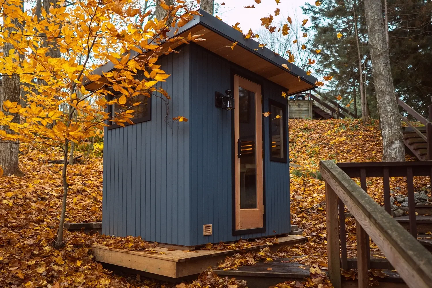 Small blue wooden cabin in fall with yellow leaves on the ground and trees surrounding it, and wooden stairs leading up to the deck.