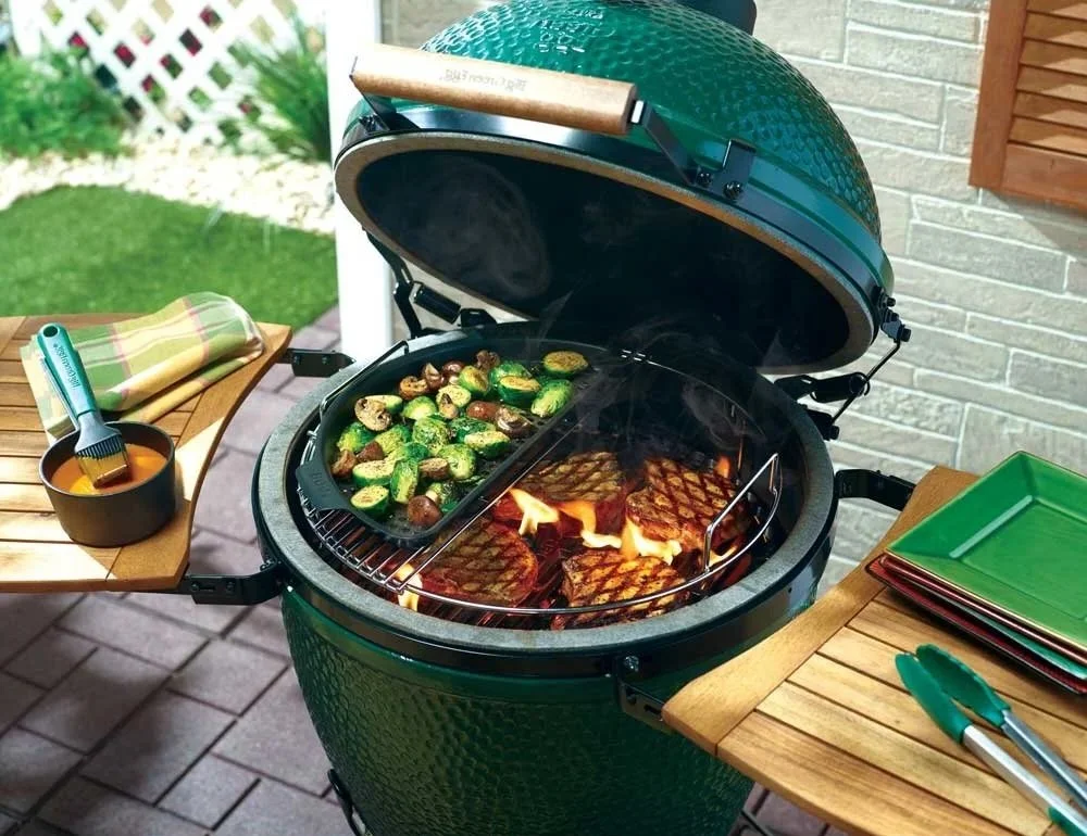 A green outdoor grill with a lid open, cooking meat and vegetables. On the left side of the grill, there is a grilling tray with sliced zucchini and mushrooms. Surrounding the grill are wooden tables with green plates, a pair of tongs, and a bowl with a basting brush.