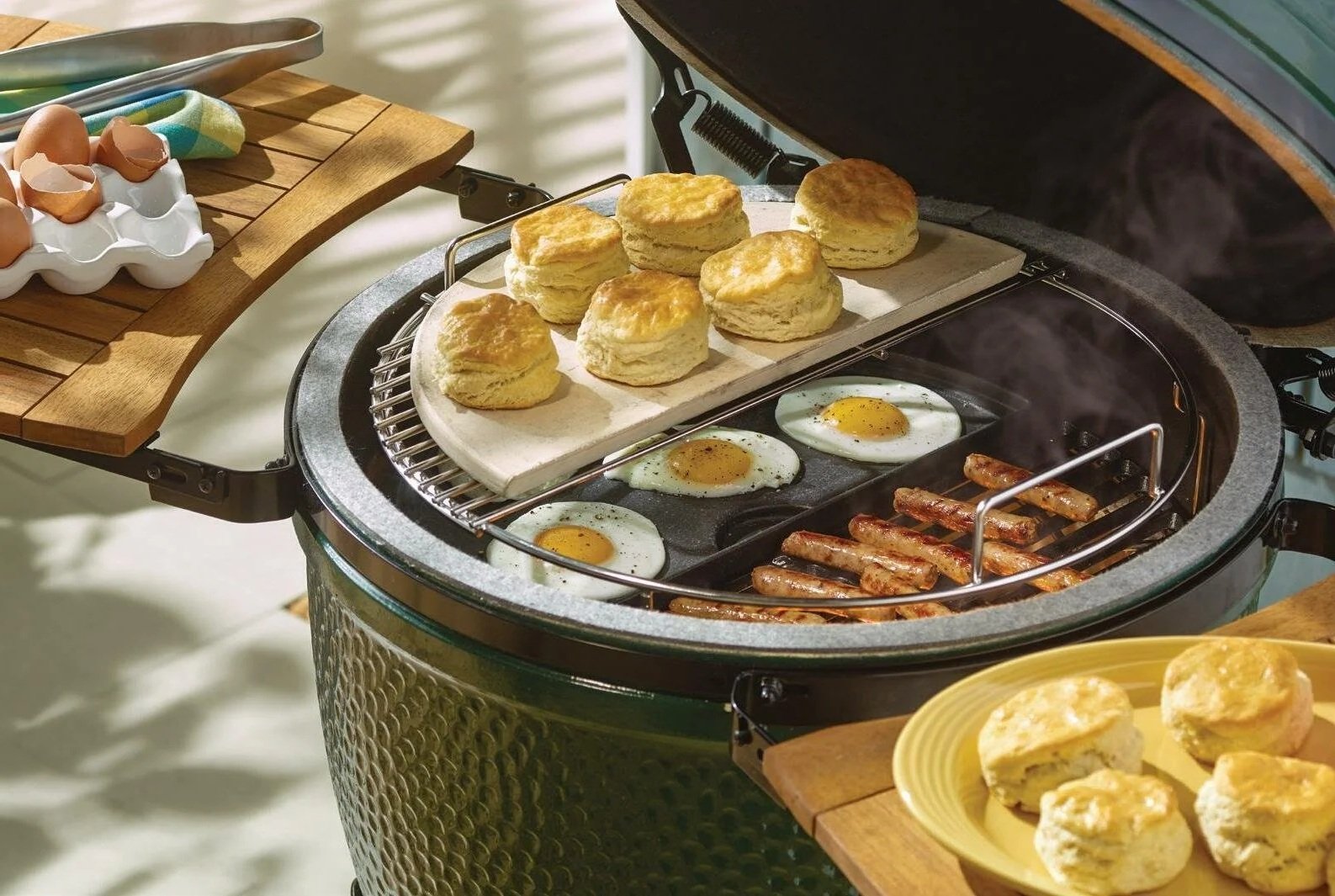 A grill with breakfast items cooking, including biscuits, fried eggs, and bacon. There are plates and a wooden tray with eggs and bread surrounding the grill.