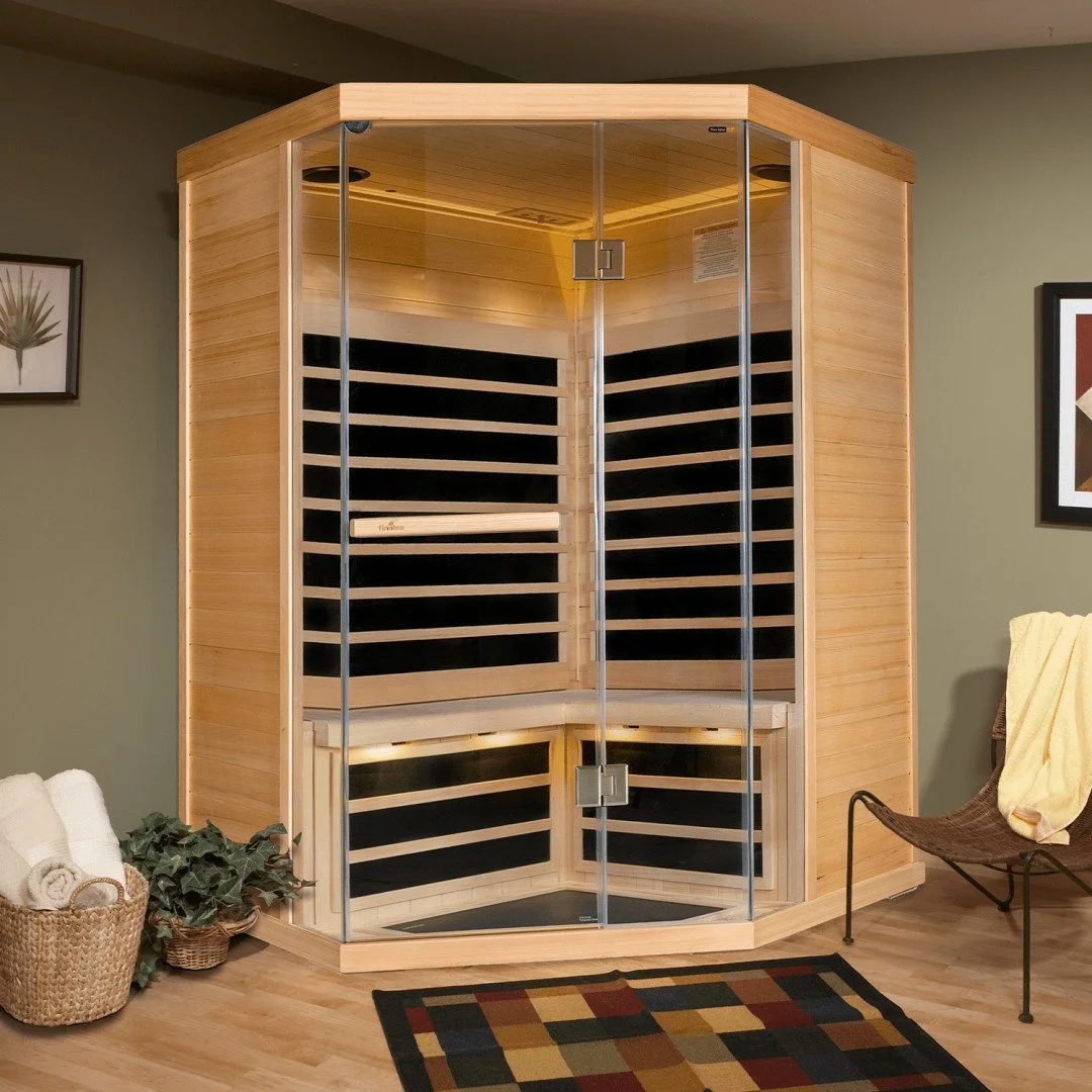 A wooden infrared sauna with glass doors in a home setting, next to a chair with a yellow towel, a houseplant, and a basket with rolled towels.
