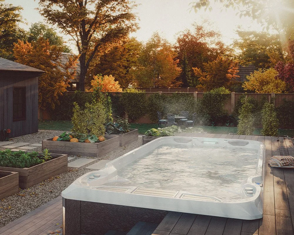 A backyard with a hot tub on a wooden deck, surrounded by garden beds filled with plants and pumpkins, with trees displaying autumn foliage in the background during sunset.