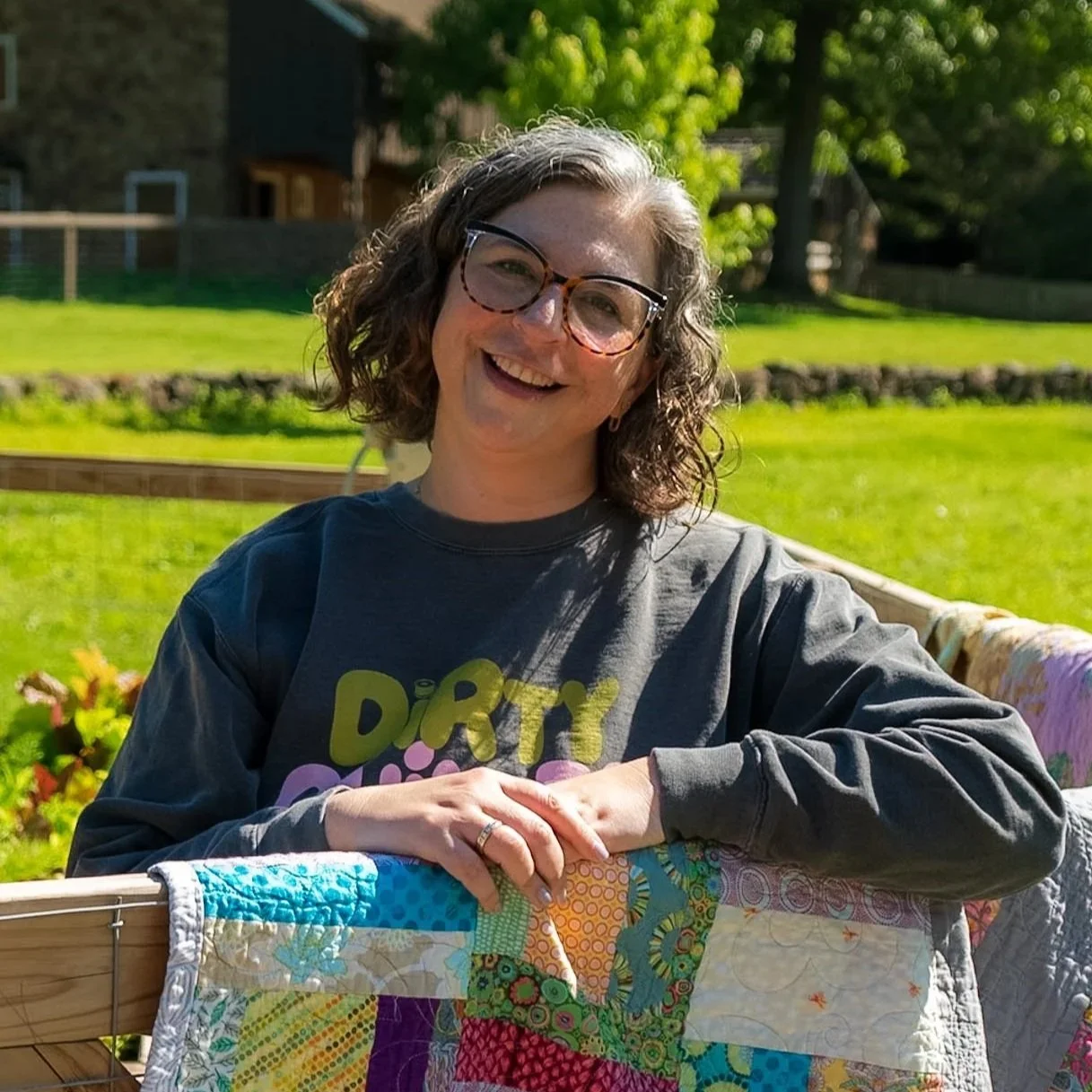 A woman with curly gray hair and glasses smiling outdoors on a sunny day, wearing a dark sweatshirt with colorful text, leaning on a quilted blanket draped over a wooden railing in a garden with green grass, trees, and a house in the background.