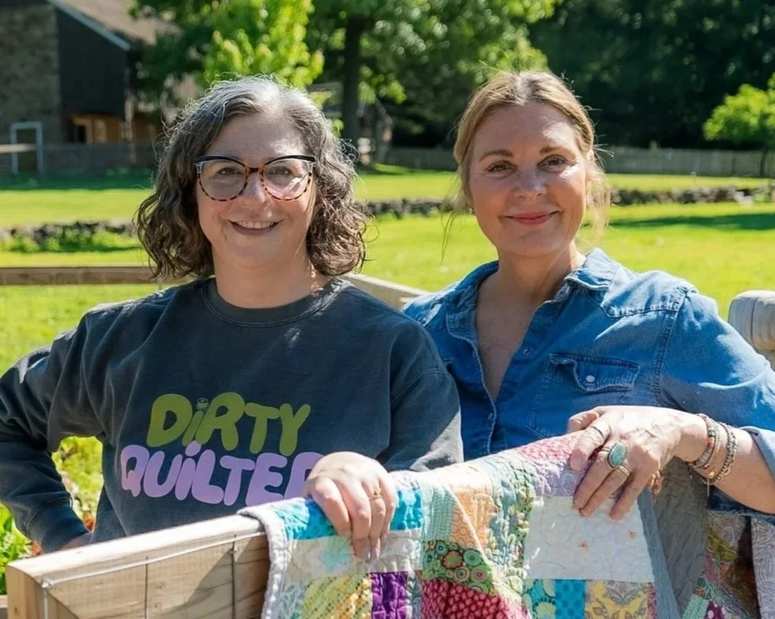 Two women smiling outdoors with greenery in the background. One woman has curly hair and glasses, wearing a dark shirt that says 'Dirty Quilter'. The other woman has blonde hair, wearing a blue denim shirt, holding a colorful quilt.