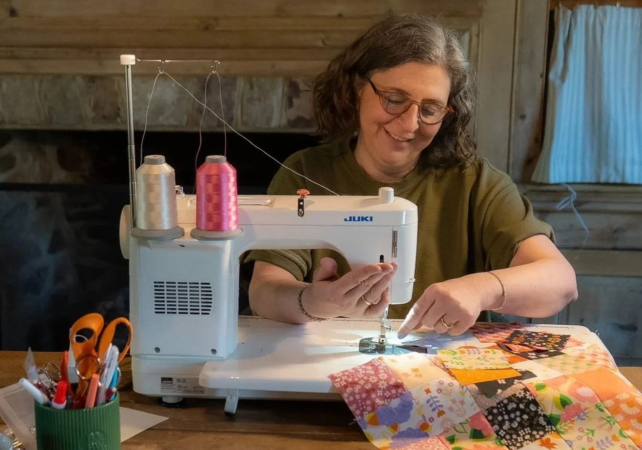 A woman sewing on a Juki sewing machine surrounded by colorful fabric and sewing supplies, in a cozy wood-paneled room.
