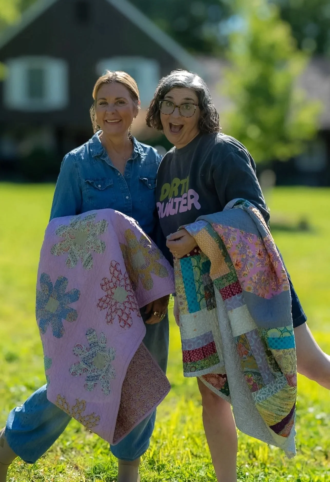 Two women smiling outdoors, holding colorful quilt blocks, with a house and trees in the background.