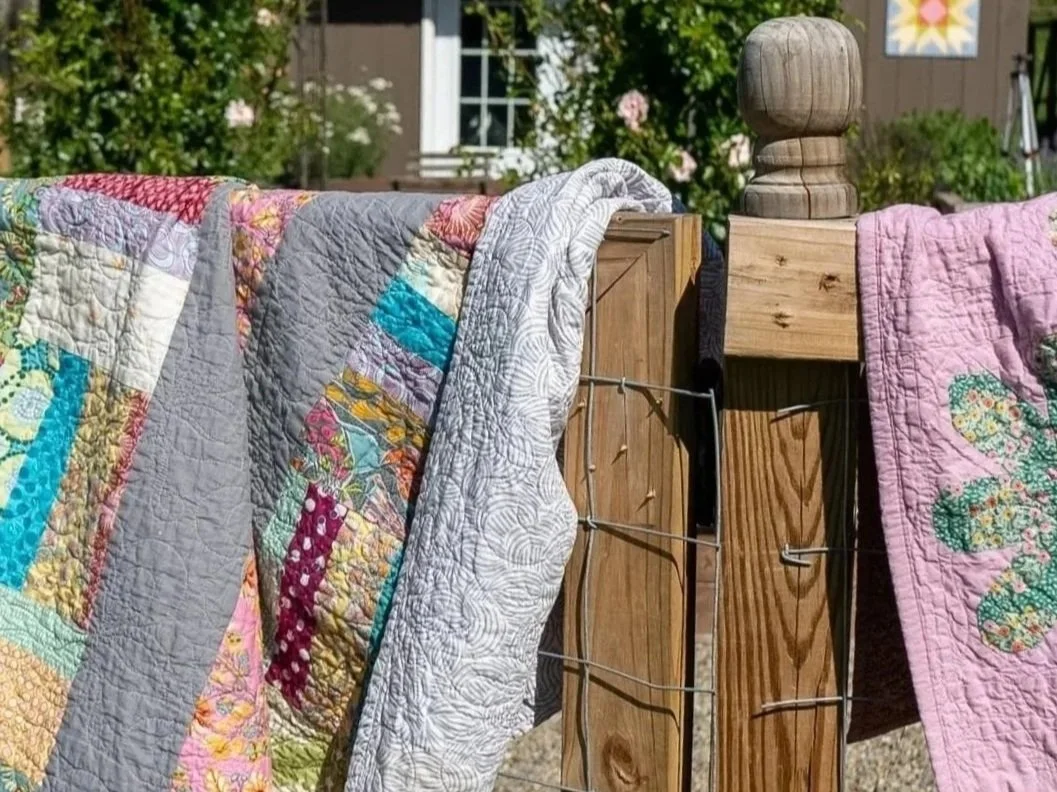 Colorful patchwork quilts hanging over a wooden fence on a sunny day.