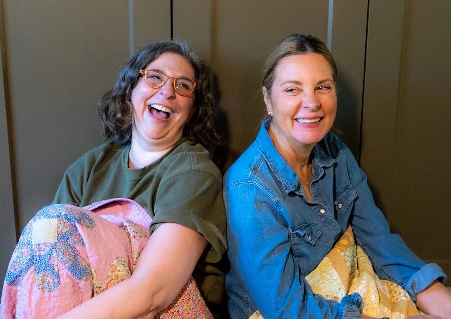 Two women sitting back to back, smiling and laughing, with one wearing glasses and a green shirt, and the other in a denim shirt, against a wooden background.