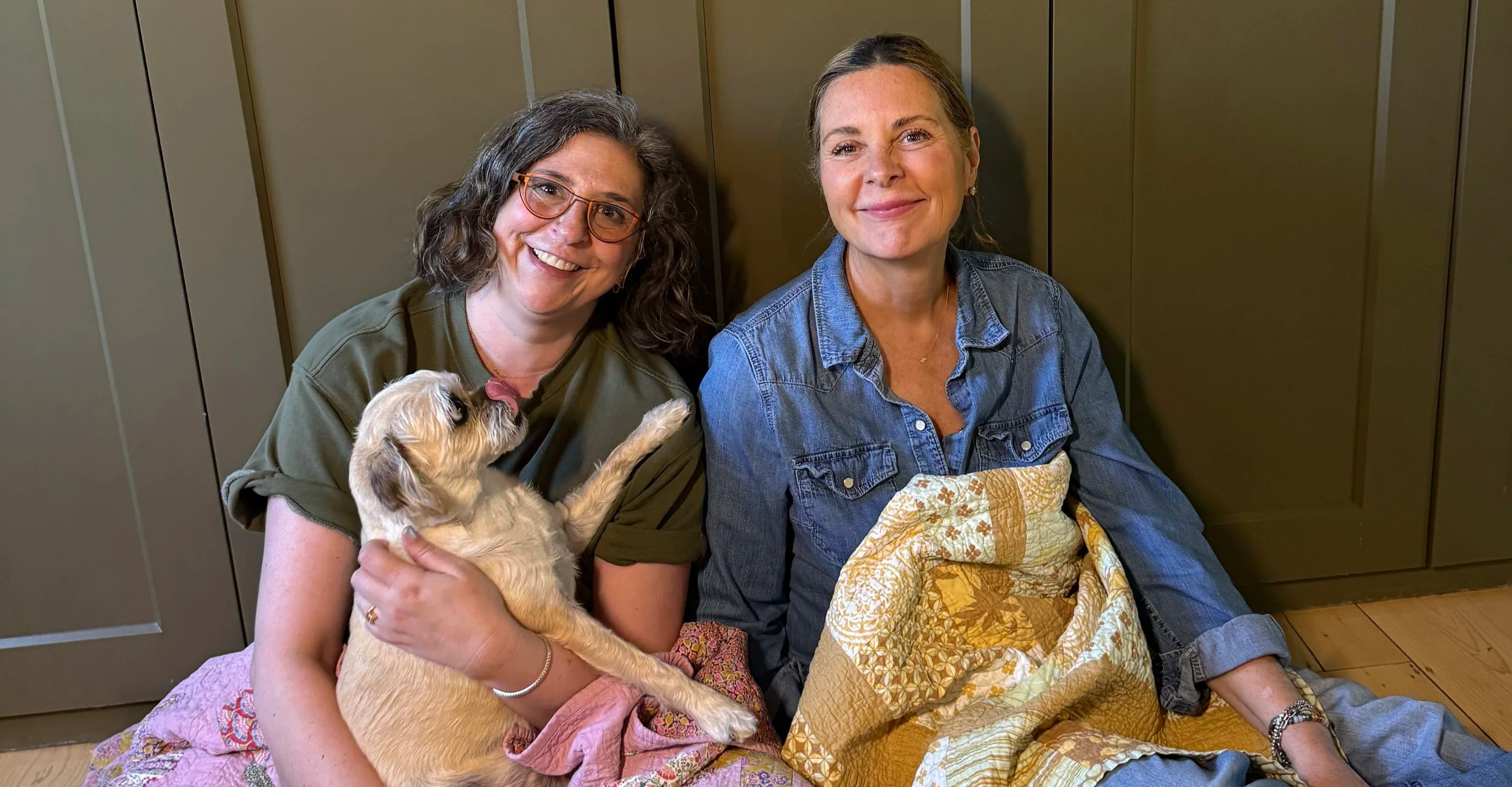 Two women sitting on the floor in front of a pantry, one holding a puppy, both smiling at the camera.