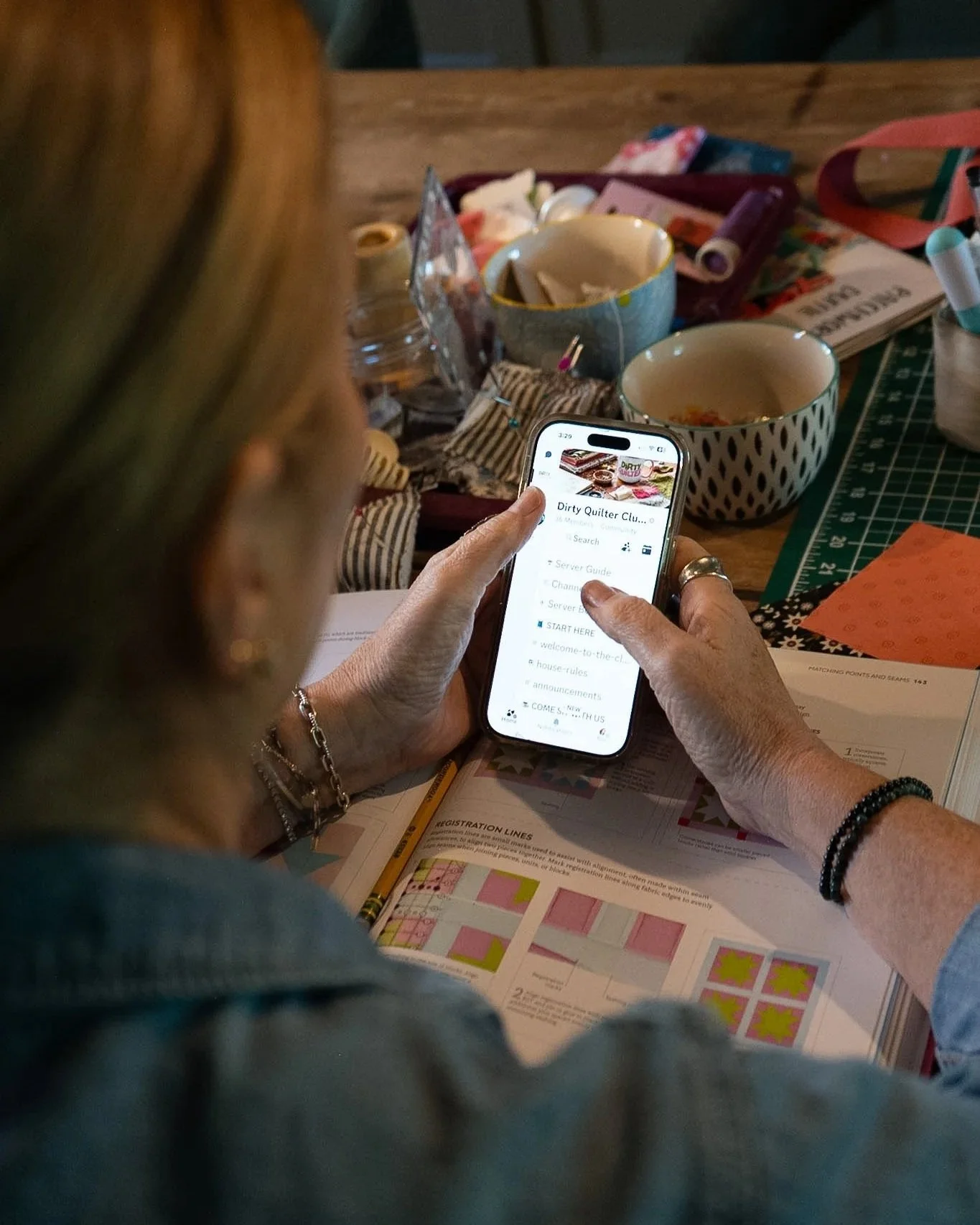 Person with blonde hair, wearing bracelets, holding a smartphone and looking at a quilting club webpage, with quilting patterns and papers spread on the table, and various sewing supplies in the background.