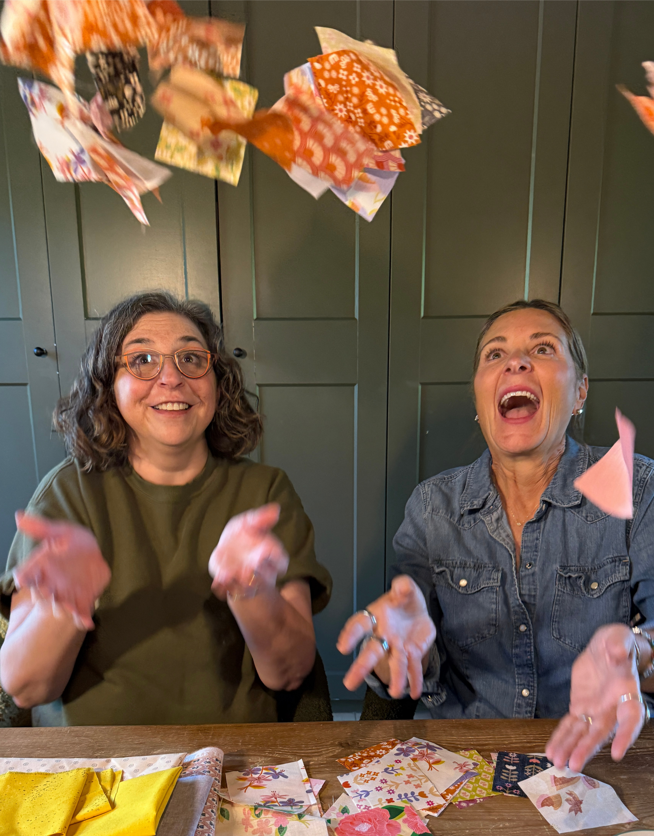 Two women are sitting at a table with colorful fabric and paper scraps, tossing pieces of fabric into the air with joyful expressions.
